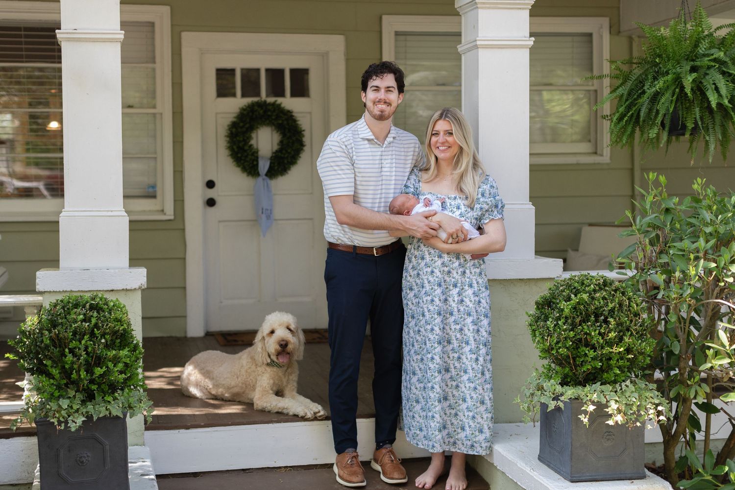 Young couple with newborn baby and golden dog pose on front porch of green home for family portrait.