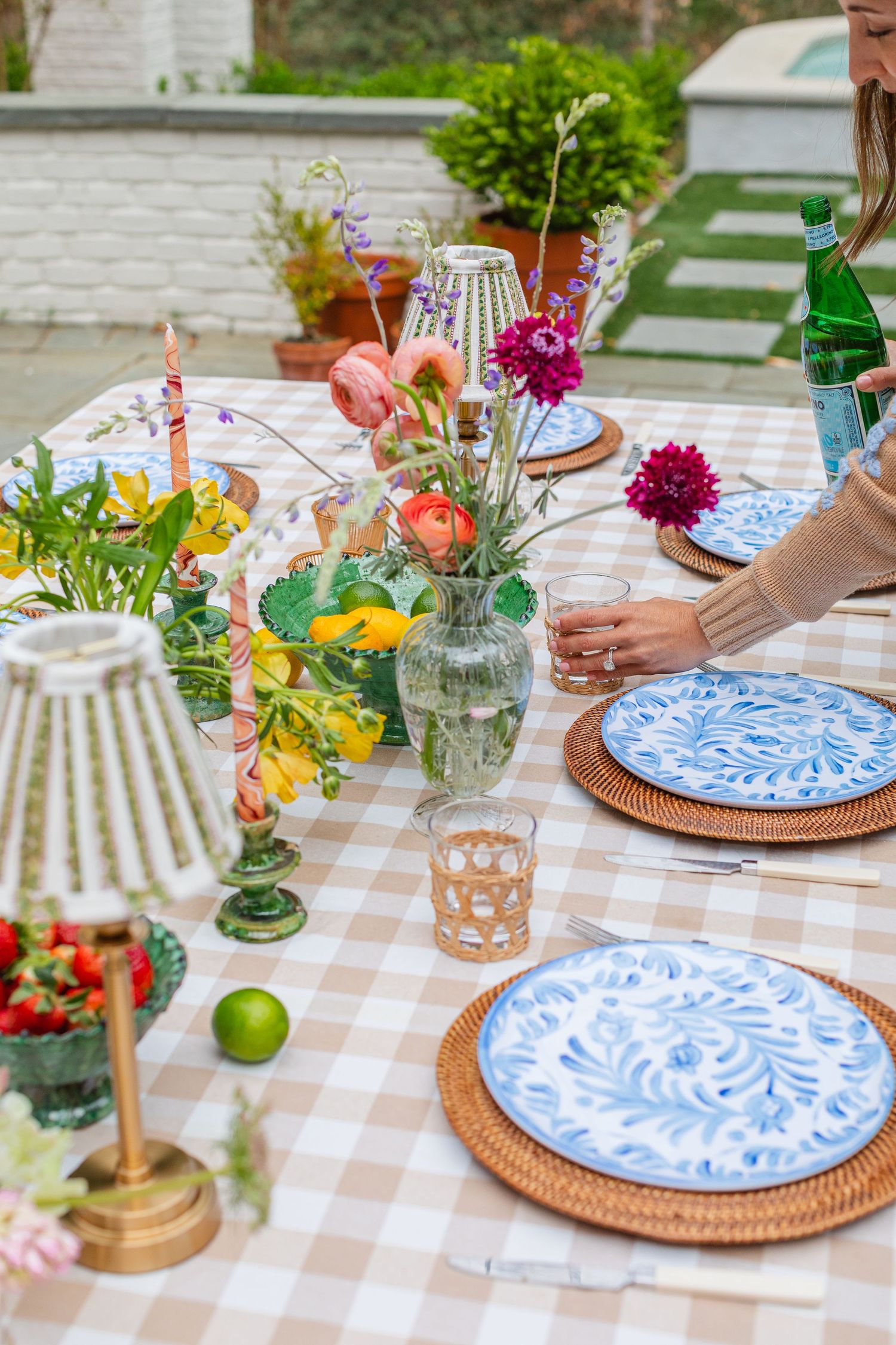 Elegant outdoor dining table with blue floral plates, rattan chargers, fresh flowers, candles, and a gingham tablecloth.
