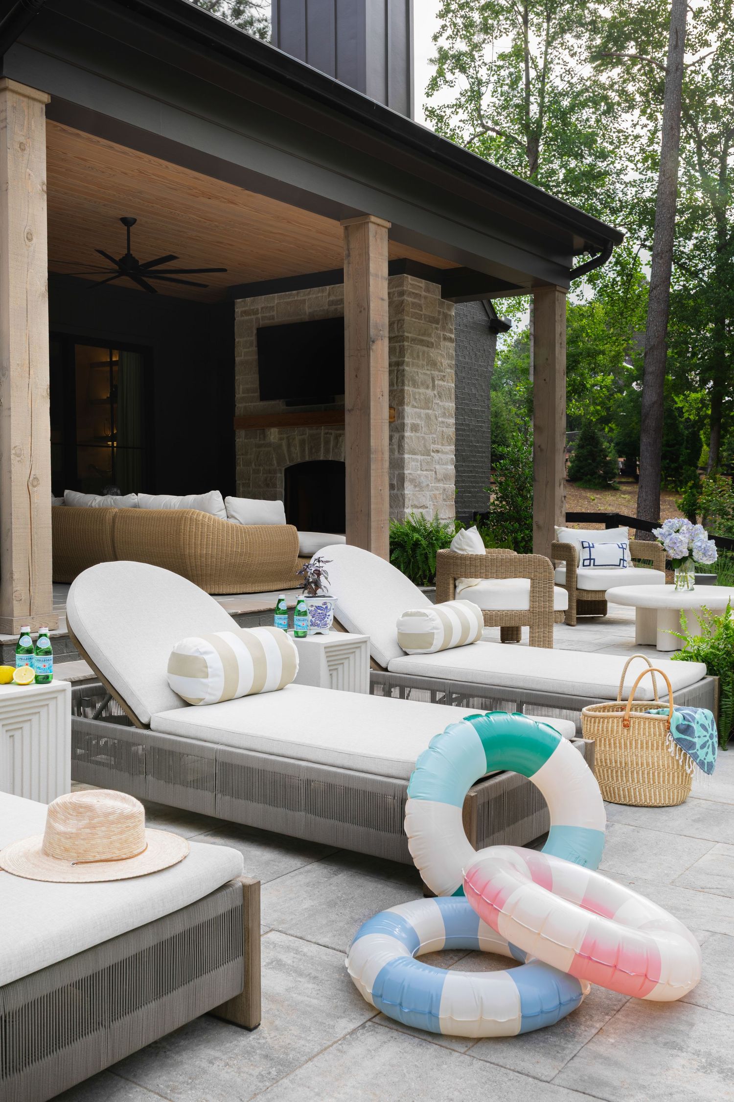 Outdoor patio with lounge chairs, pool floats, wicker furniture, covered pergola with fireplace, and lush green trees.