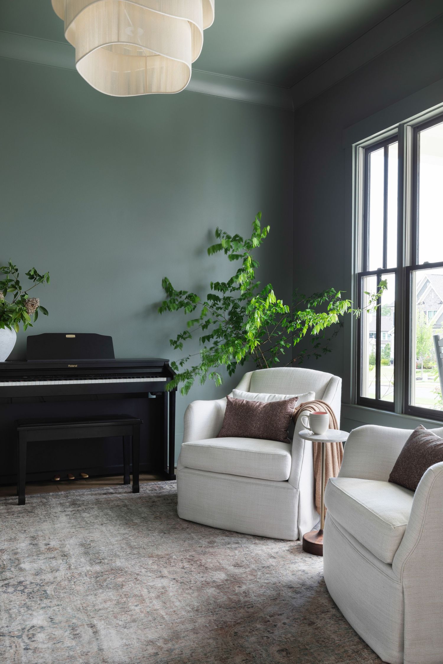 Elegant sitting room with sage green walls, white armchairs, a black digital piano, and a tall green plant by dark-framed windows.