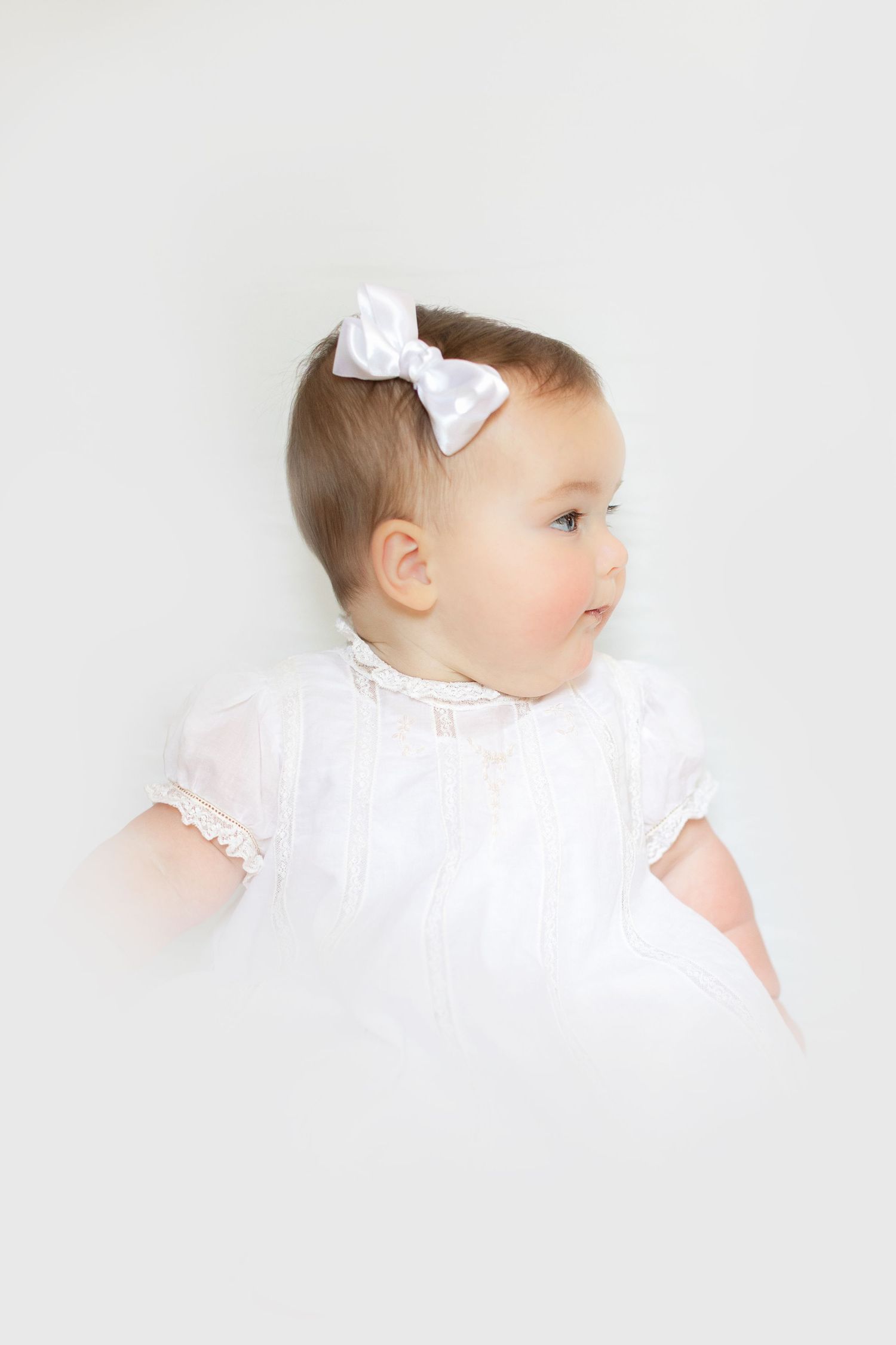 Adorable baby girl in white lace dress with satin bow headband looking sideways against white background.