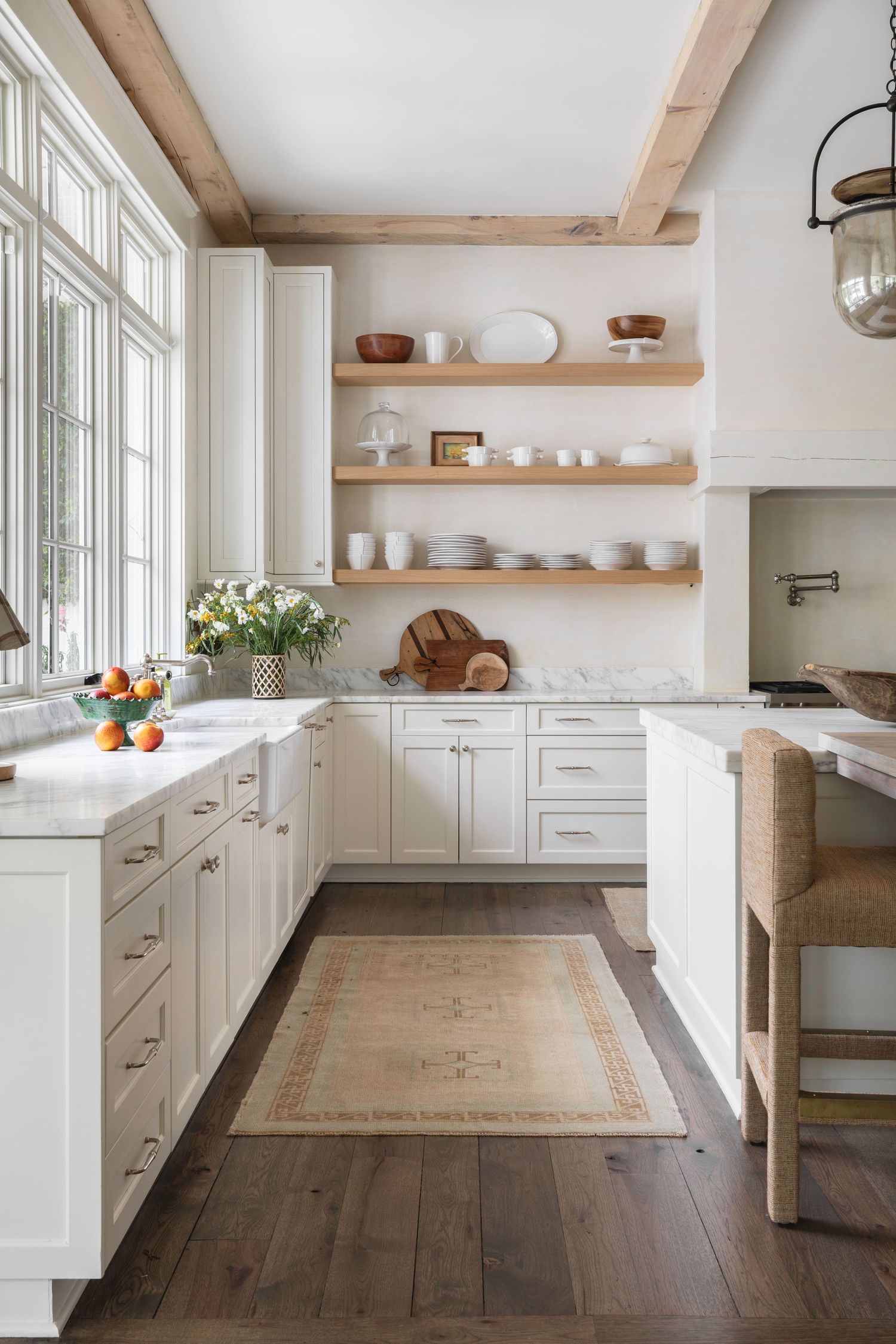 Bright white farmhouse kitchen with open wood shelves, marble countertops, dark hardwood floors, and natural beam accents.