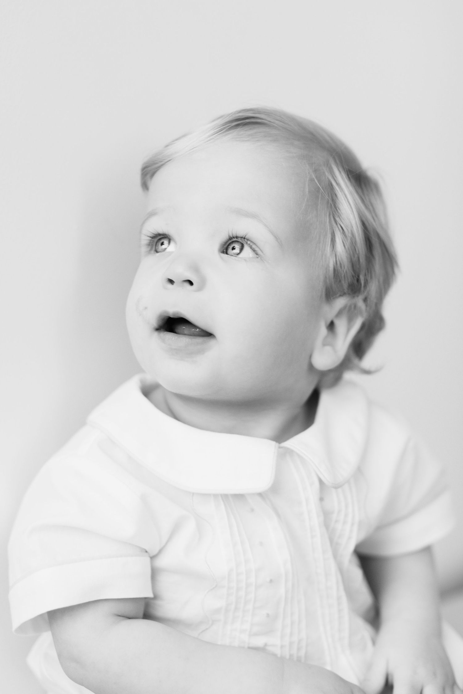High-key black and white portraits of baby in white outfit looking upward.