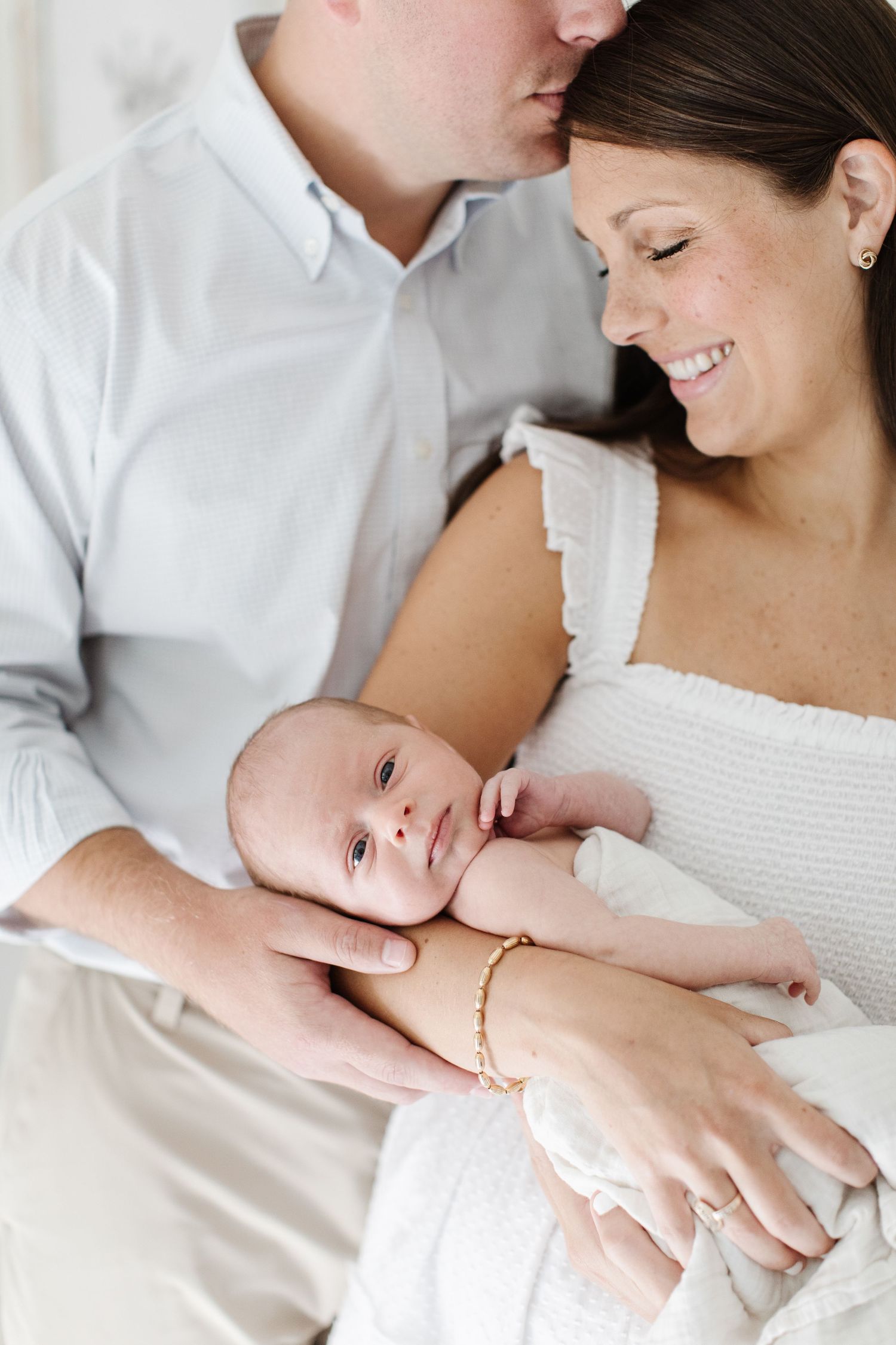Parents in white and gray outfits share tender moments with newborn baby in bright indoor setting.
