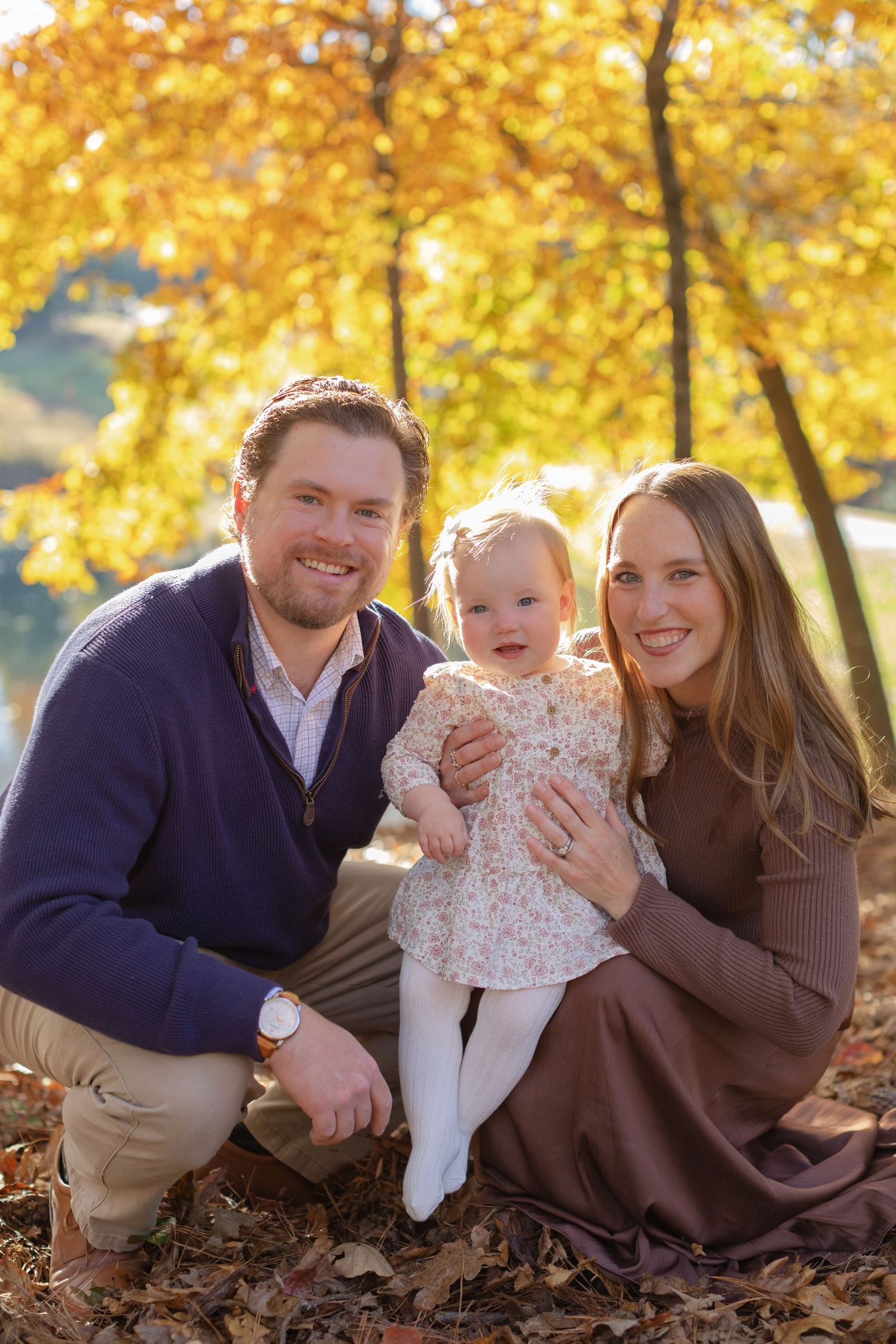 Family sitting together under golden autumn trees with fallen leaves during outdoor photo session.