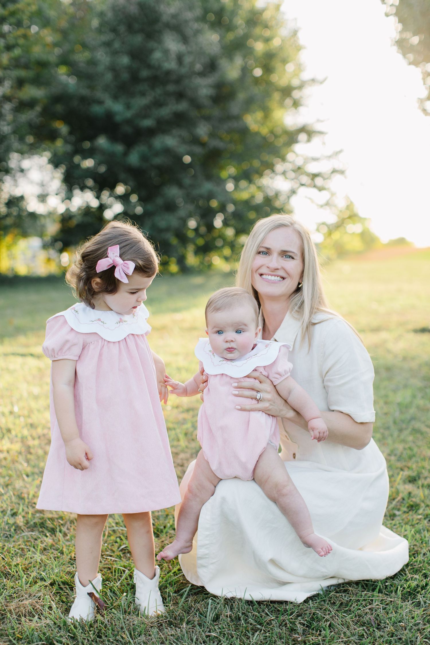 A tender outdoor family photoshoot in soft pink and white outfits during golden hour in a grassy field.