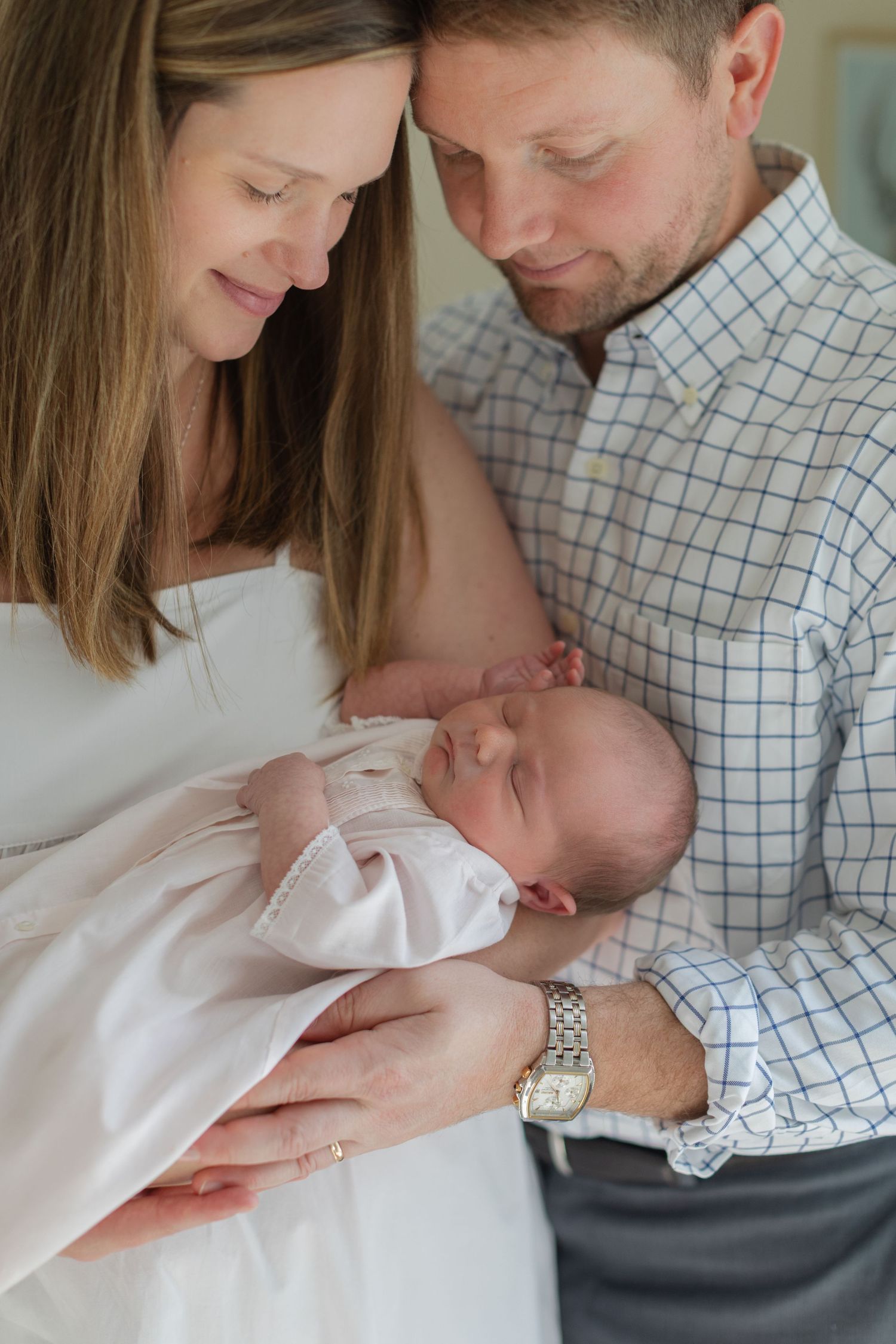 Parents in matching white and checkered outfits tenderly hold their newborn baby in a series of intimate portraits.