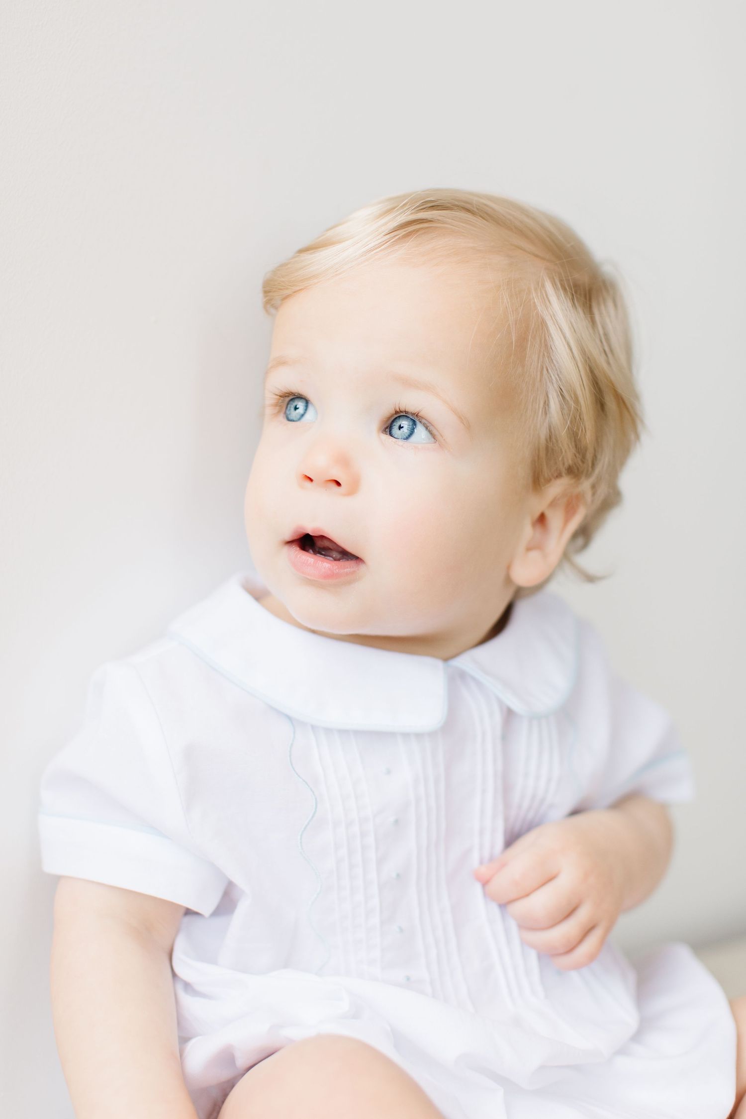 Baby in white dress with Peter Pan collar gazes upward with bright blue eyes against light background.