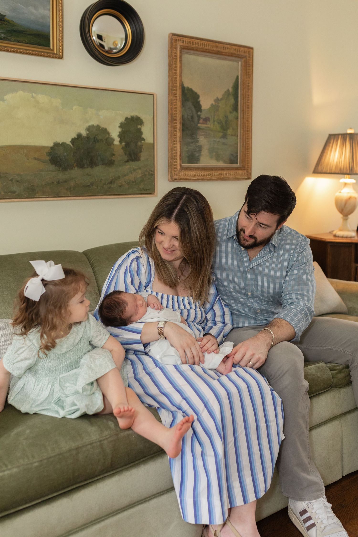 A family sits together on a green couch in a living room decorated with landscape paintings and a table lamp.