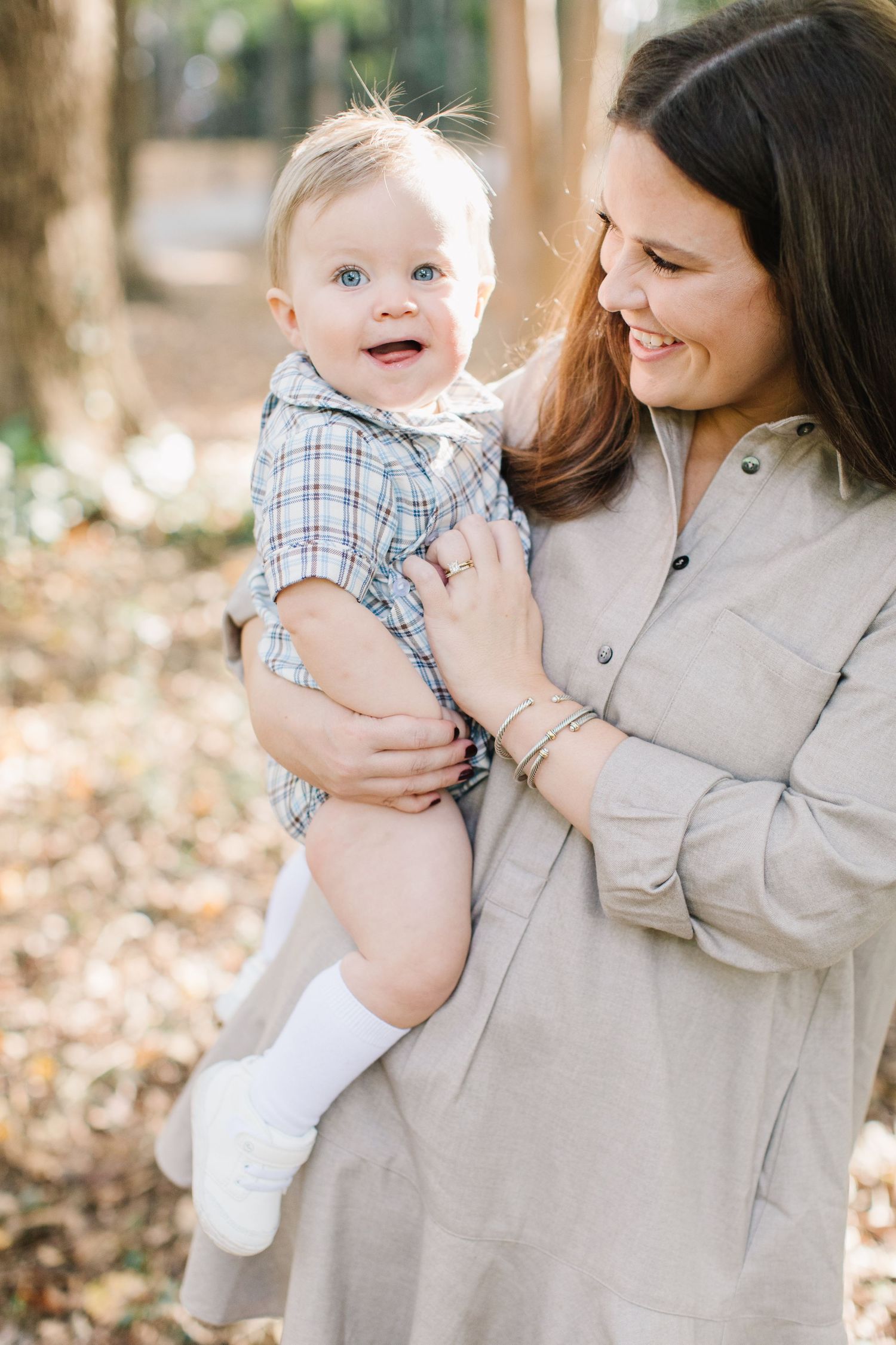 A parent in beige coat holds a laughing baby wearing plaid and white in an outdoor autumn setting.