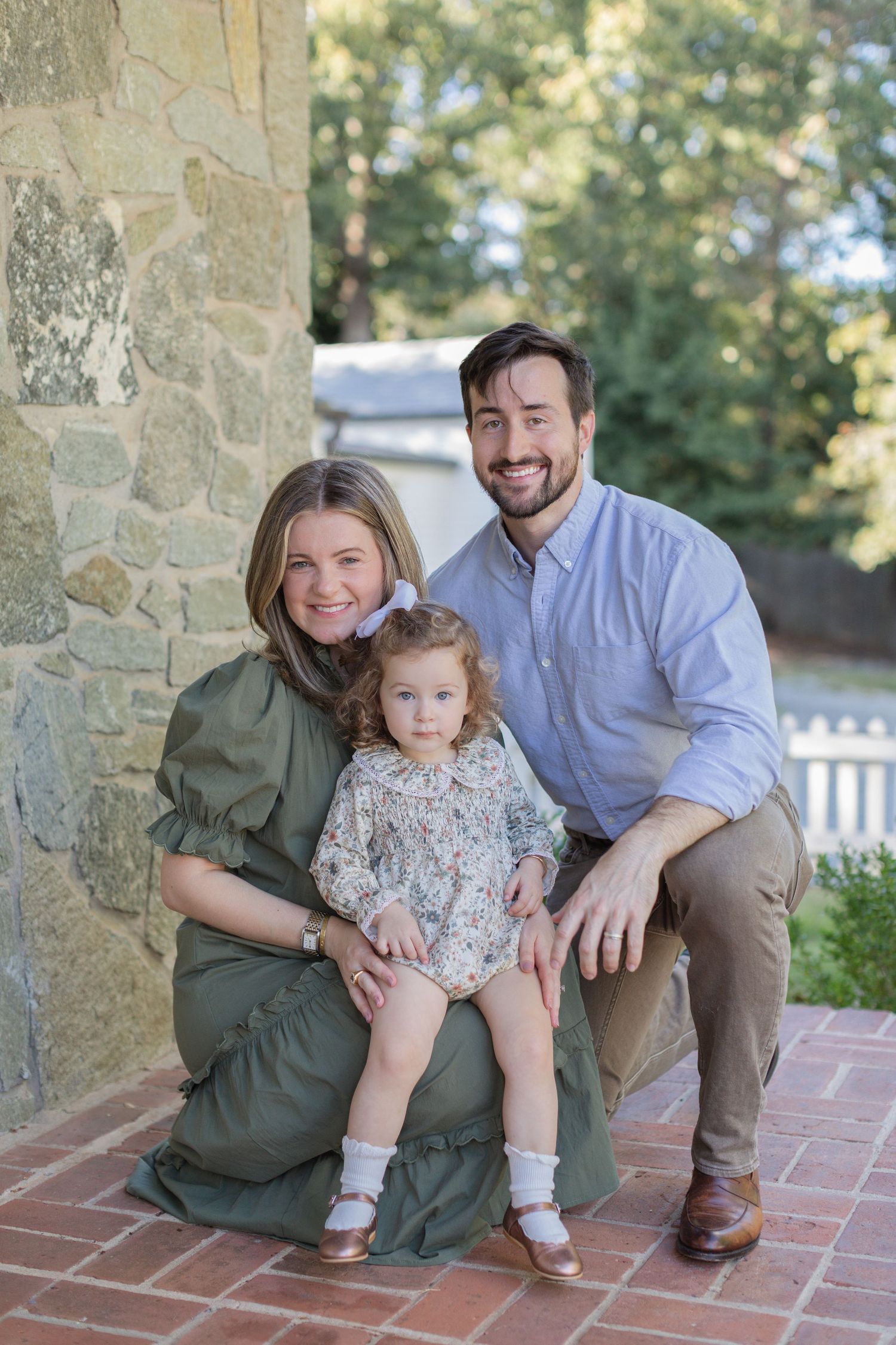 A family of three sits together on brick steps in front of a stone wall during an outdoor photo session.