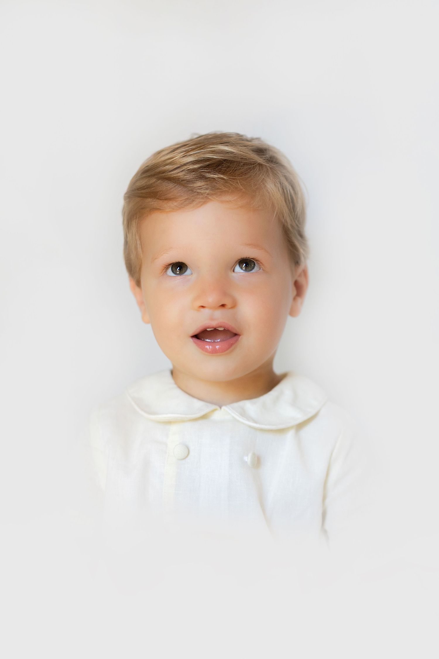 Toddler boy with blonde hair wearing a cream white Peter Pan collar outfit against a white background.