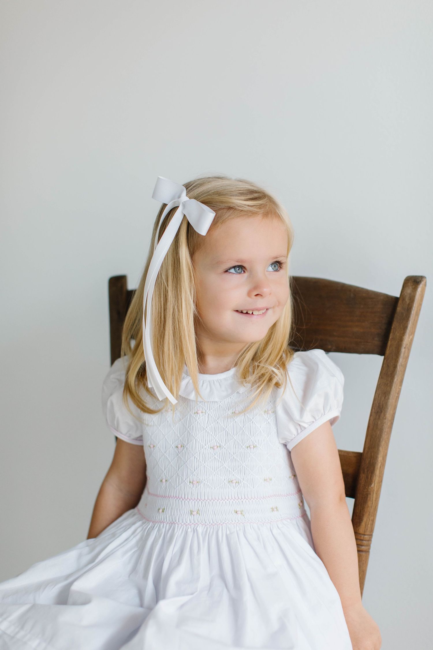 Child in white smocked dress sitting on wooden chair with white hair bow against light background.