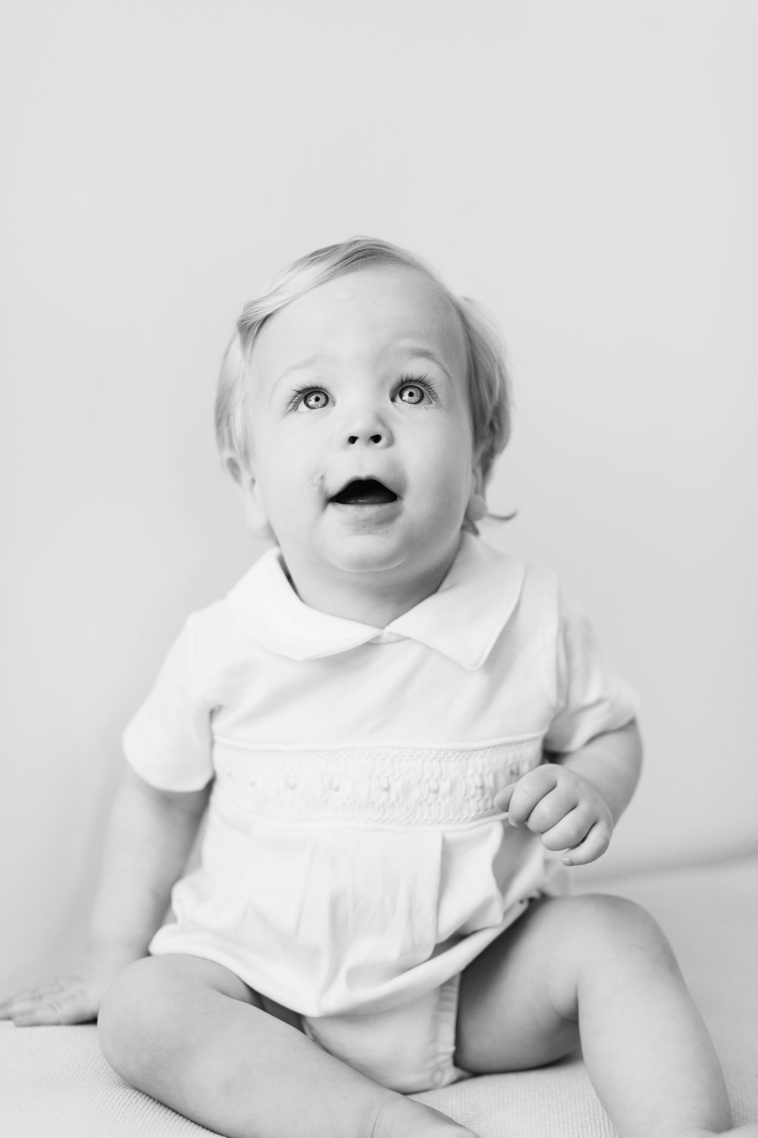Baby wearing white dress looks up with joy in minimalist black and white photo series.