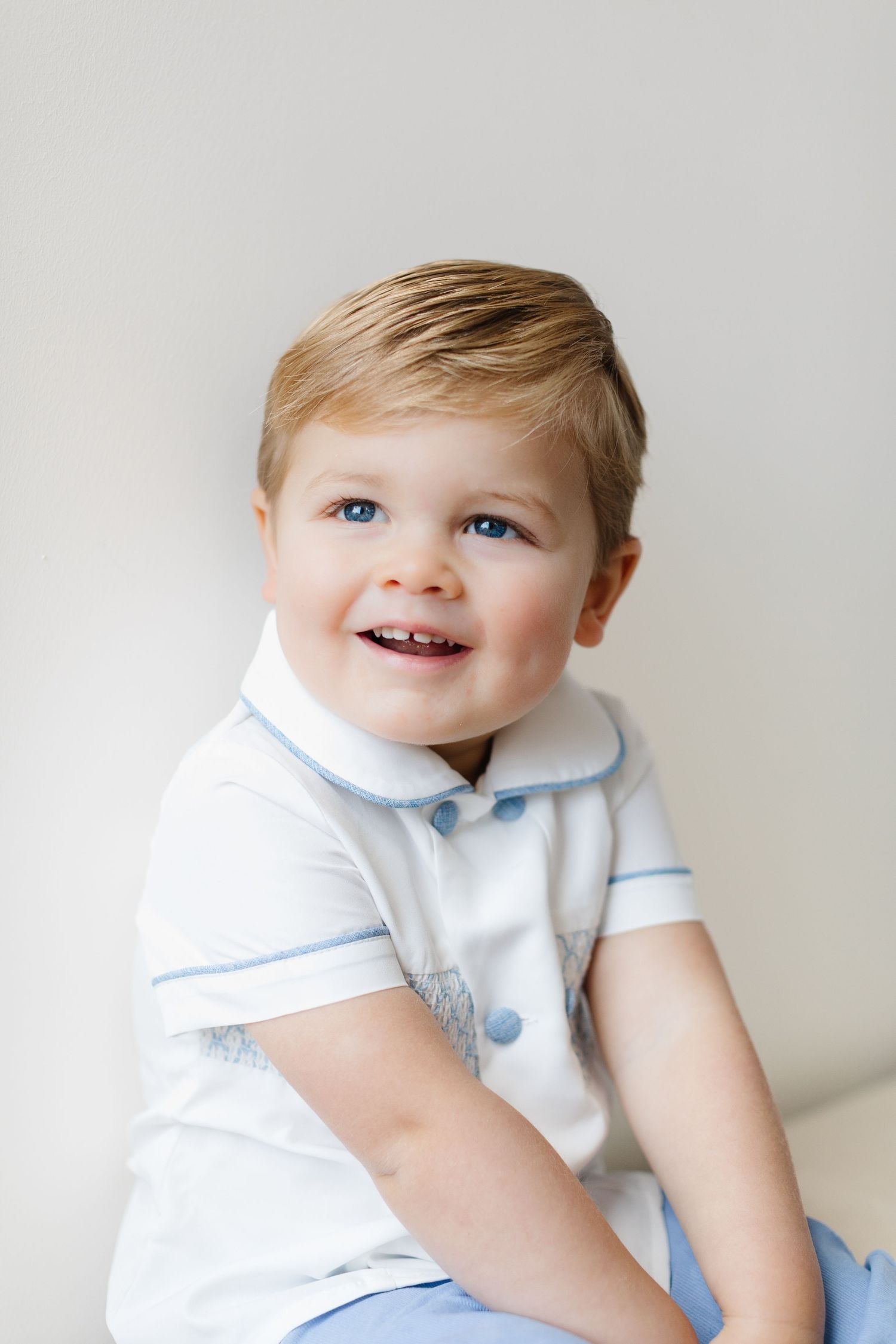 Toddler wearing a white and blue outfit smiles brightly against a light background.