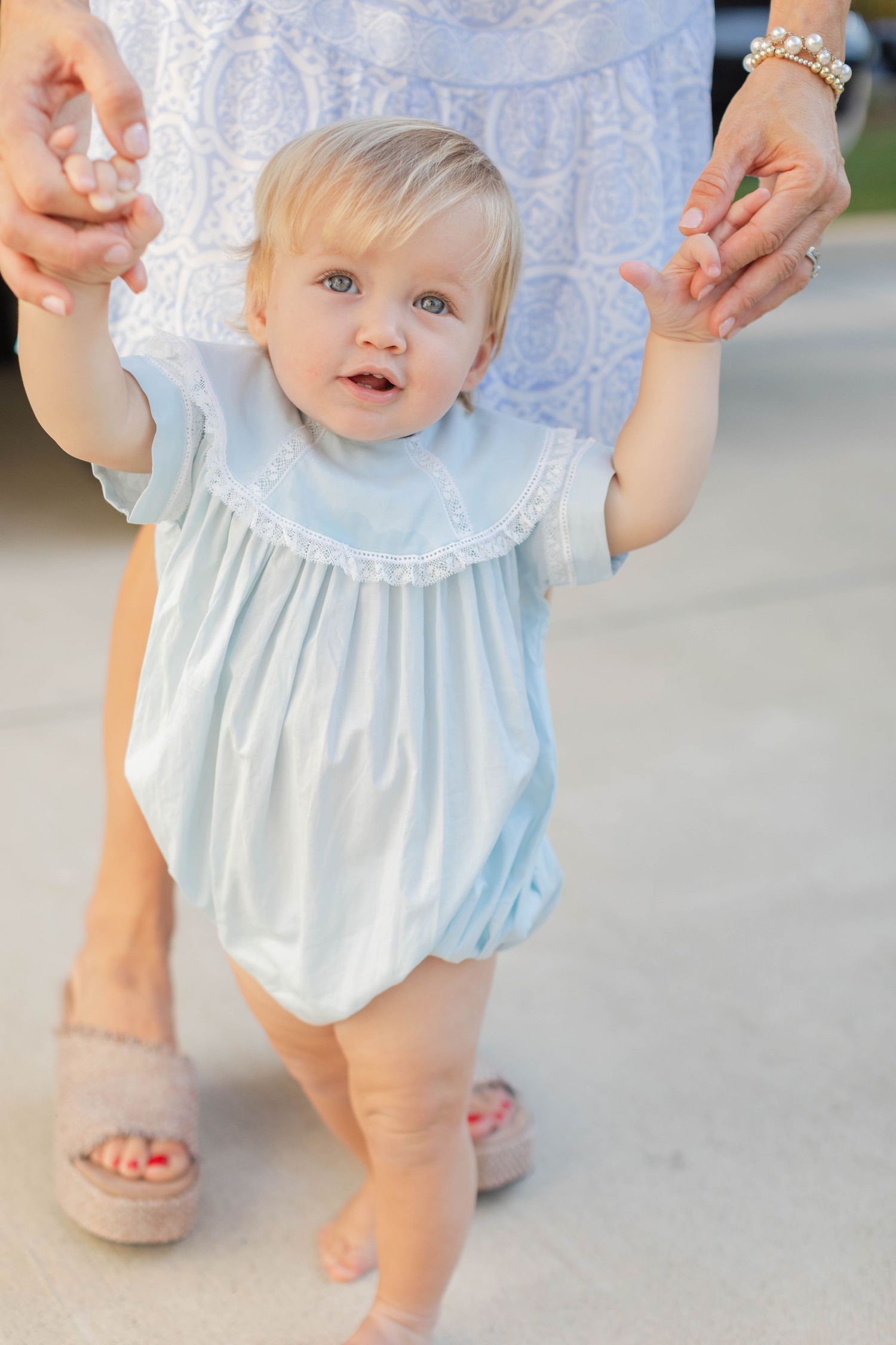 Baby in light blue dress taking first steps while holding adult hands in supportive guidance.