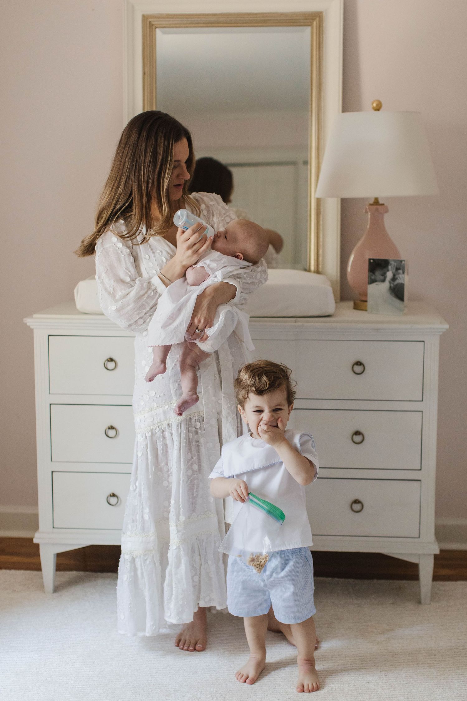 Family in white outfits bonding with newborn baby in a bright bedroom with white furniture.