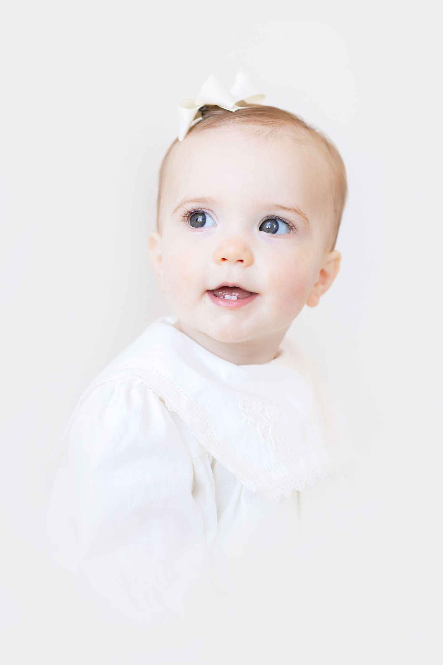 Adorable baby girl with blue eyes wearing a white dress and bow, smiling against a white background.