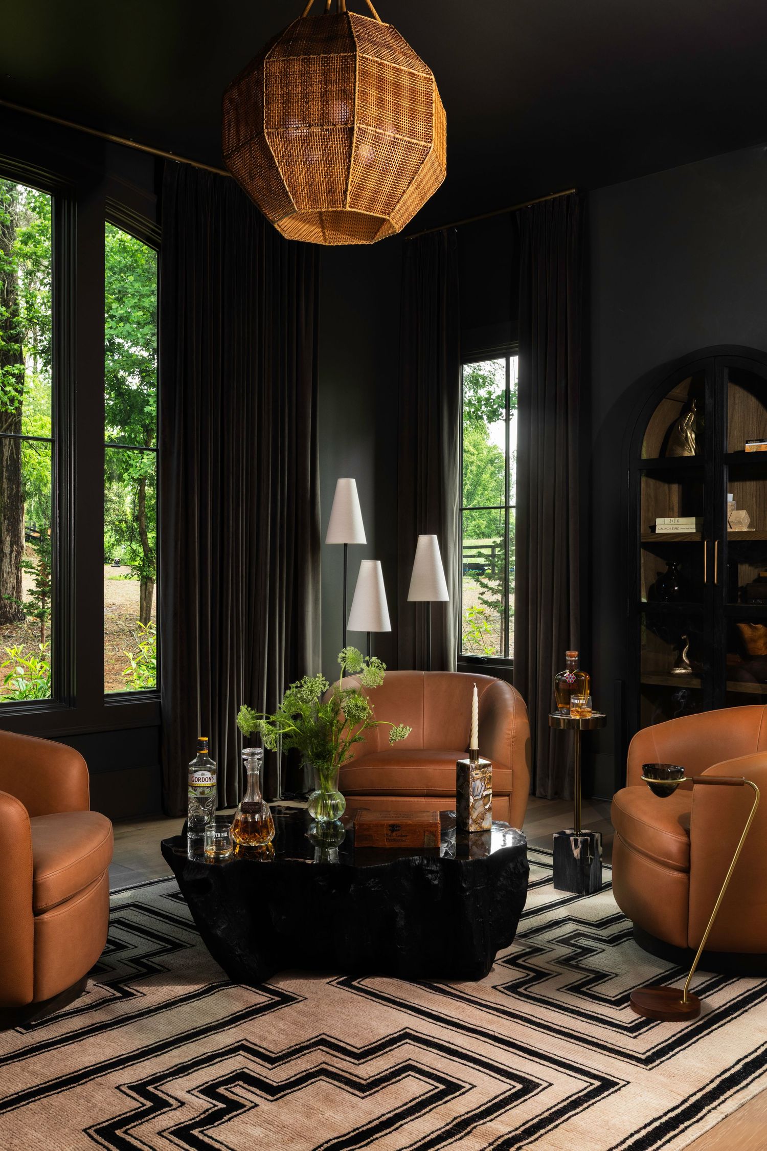 Dark moody living room with cognac leather chairs, geometric rug, black coffee table, rattan pendant light, and floor lamps.