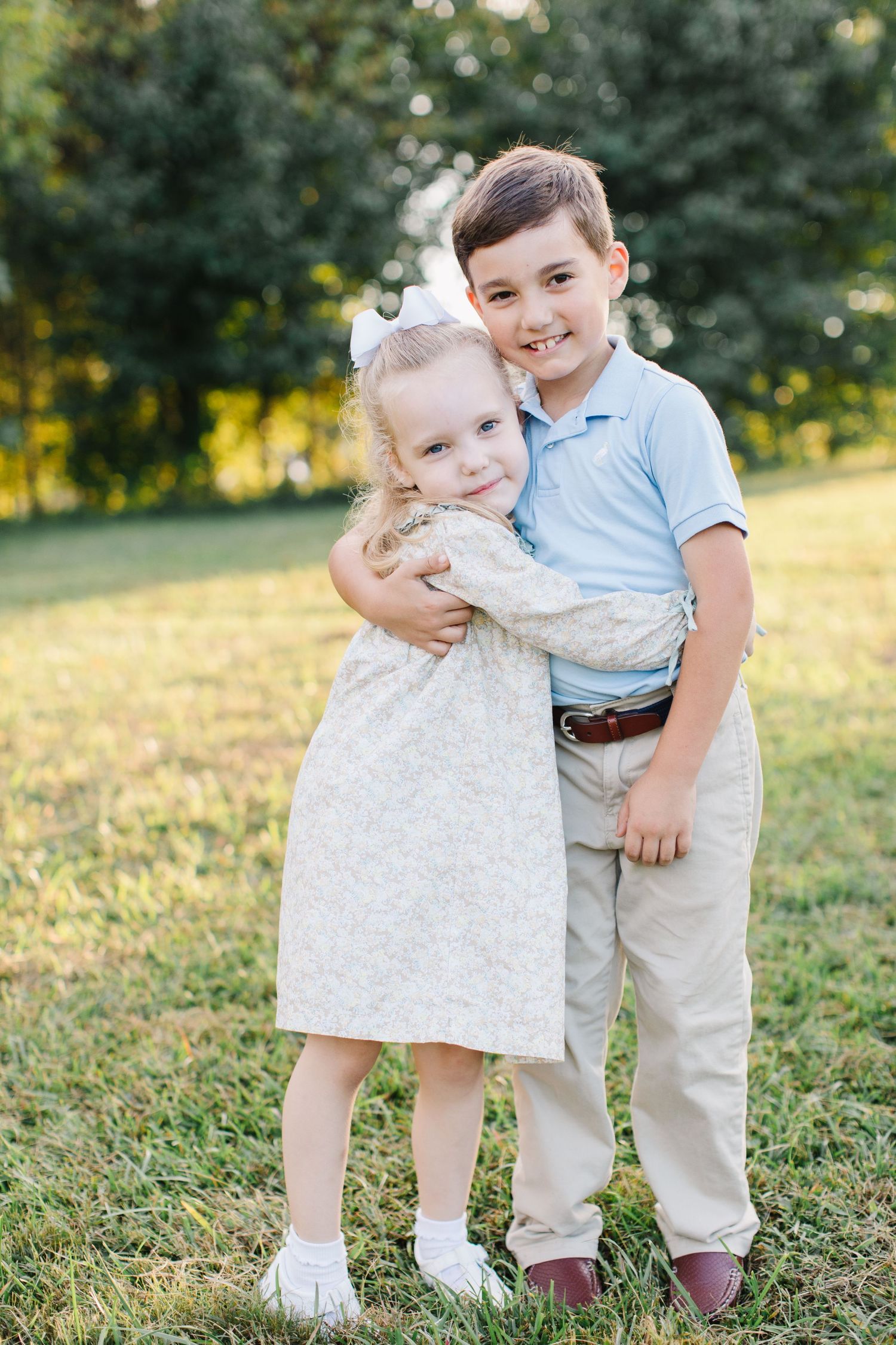 Two young children in light colored outfits embrace while standing in a grassy field at golden hour.