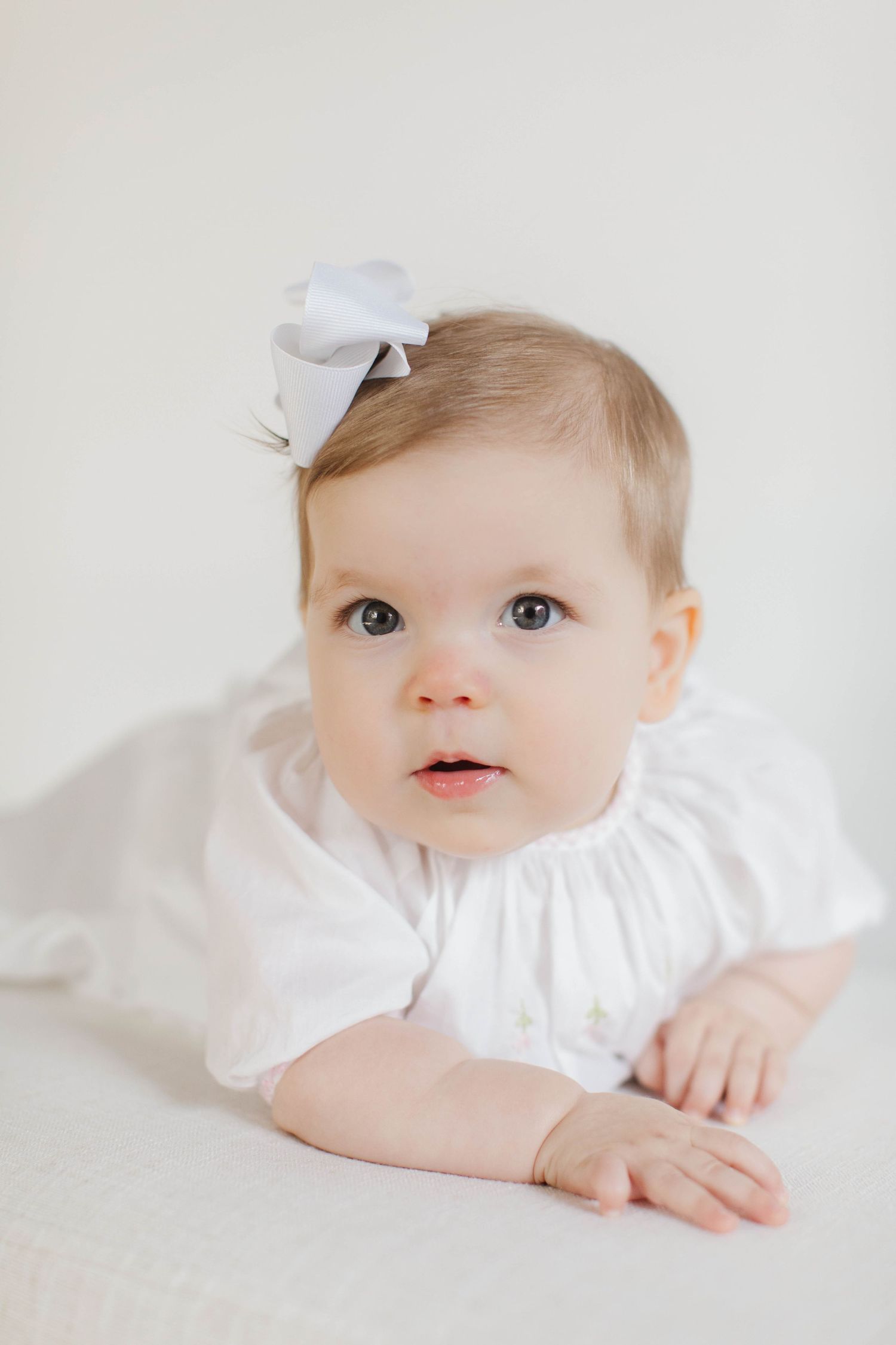 A baby wearing a white ruffle dress and bow lies on a light background looking upward with wide eyes.