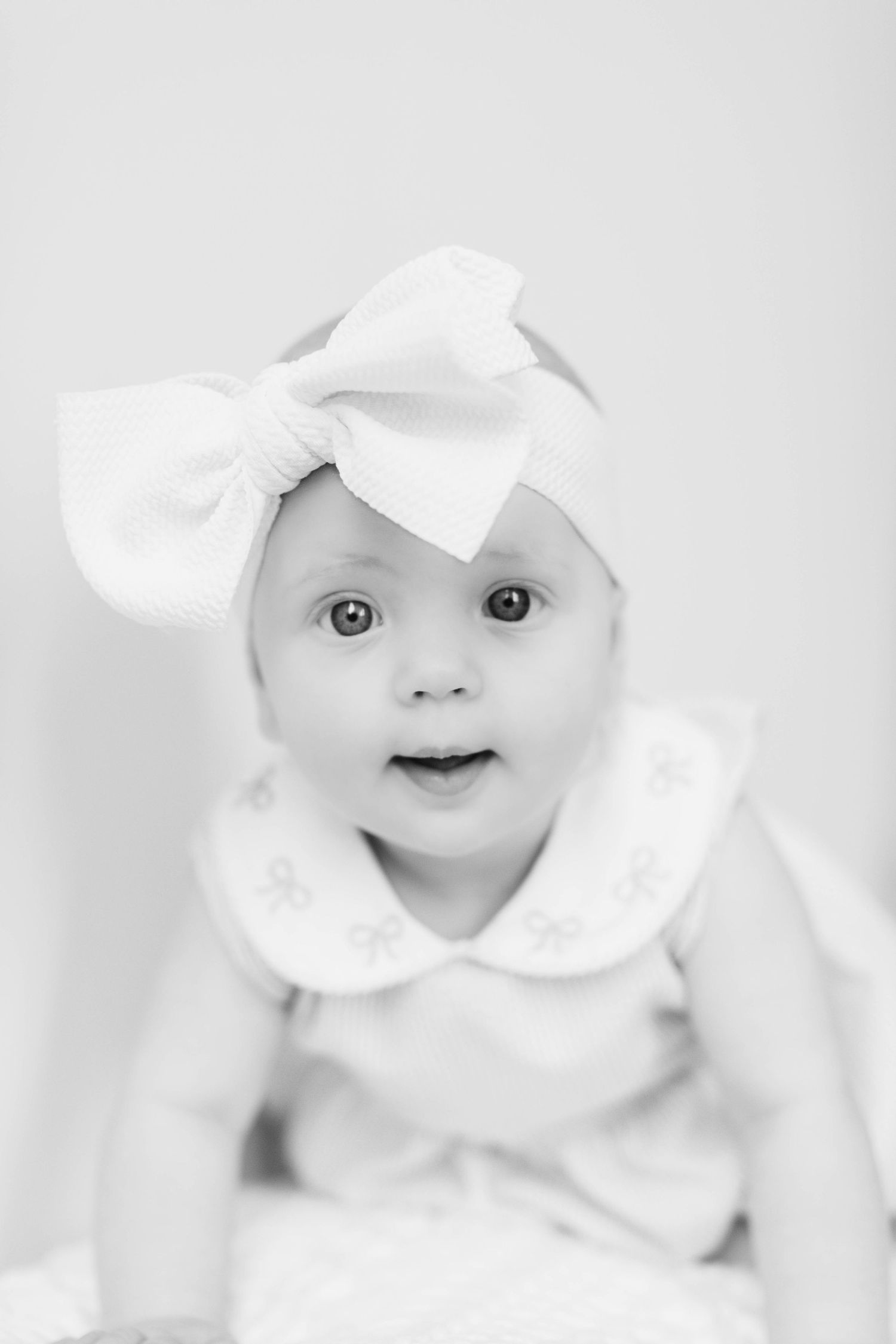 Baby in white outfit with large bow headband against light background in monochrome portrait.