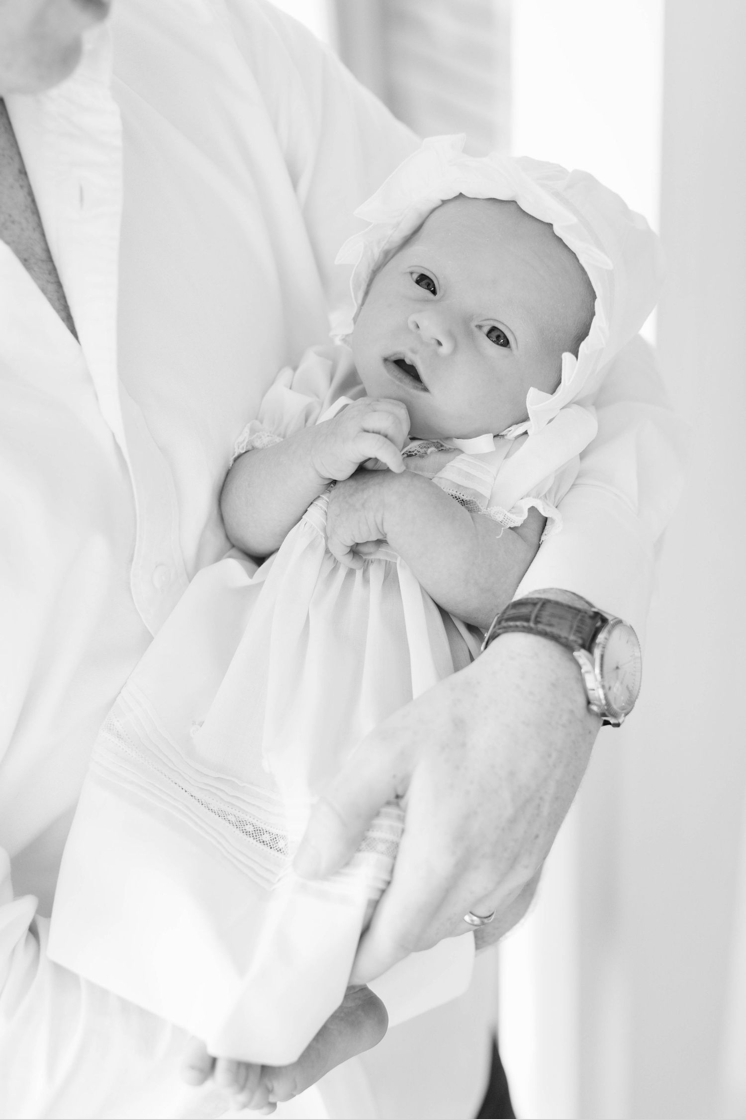 A baby wrapped in white blankets lies peacefully on a soft bed in a monochromatic high-key photograph.