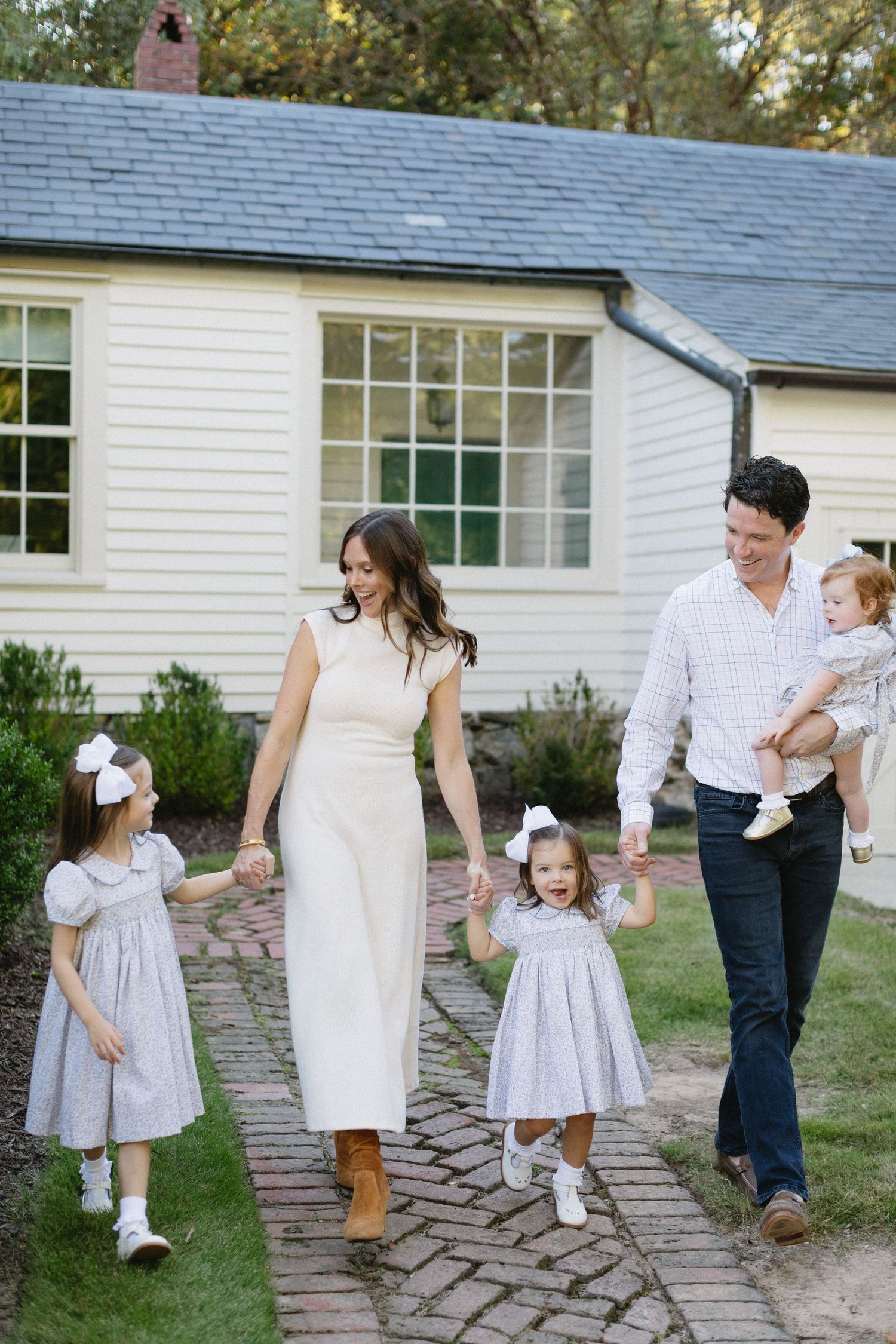 Family walks together on brick path outside white cottage house with children wearing matching gray dresses.