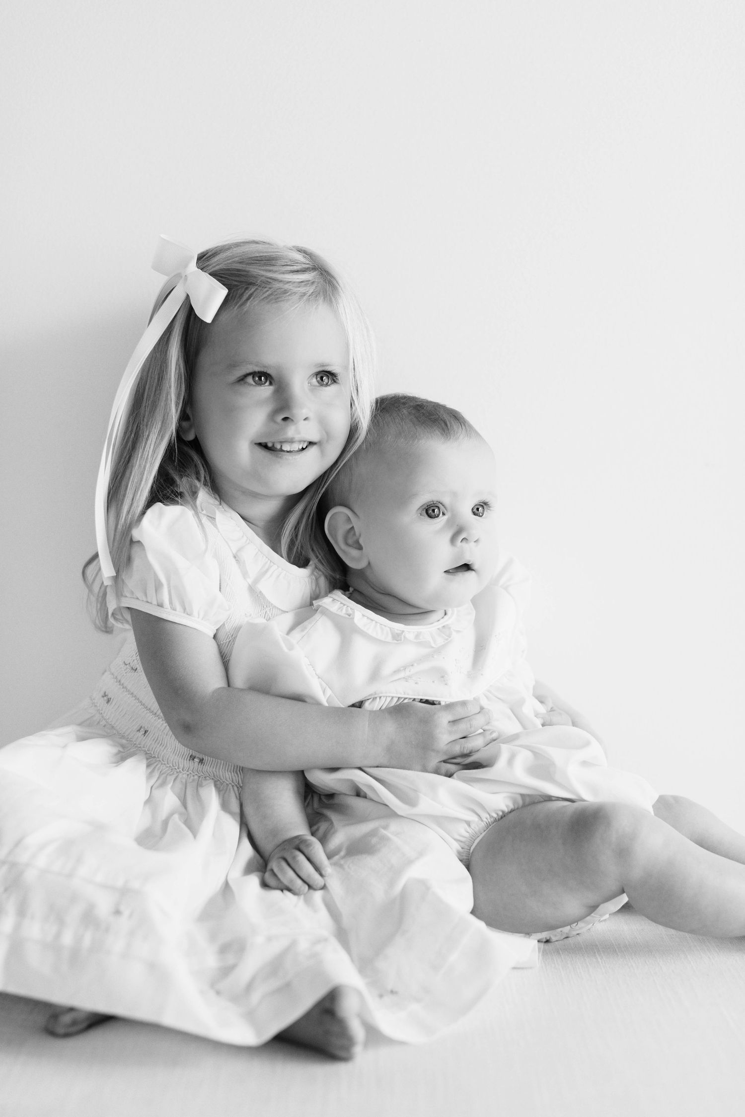 Two children in white dresses sitting together on white backdrop in elegant black and white studio photography.