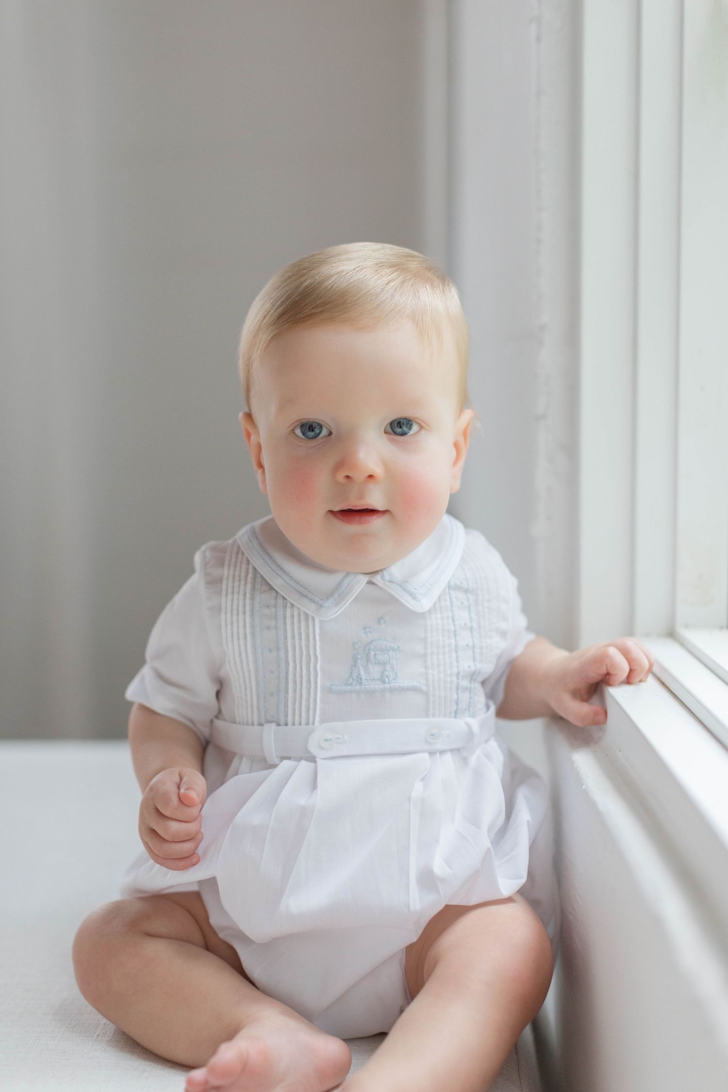 A baby in a white formal romper sits by a window in soft natural lighting.