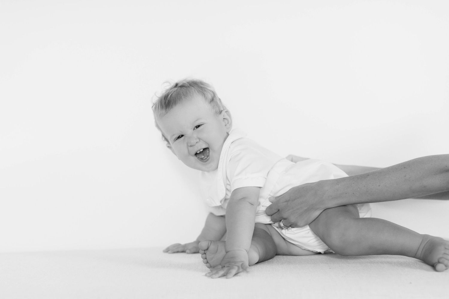 Happy infant wearing a white onesie makes playful expressions while lying on soft white bedding.