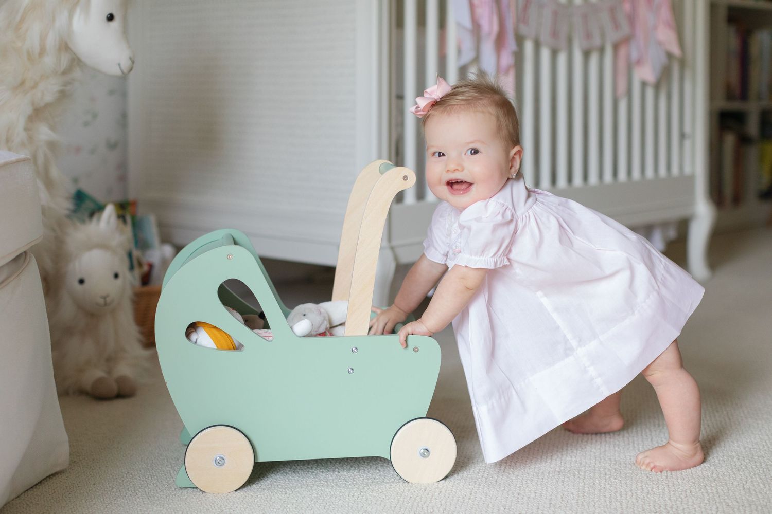 Baby girl in pink dress pushes a mint green wooden toy pram in a bright nursery room.