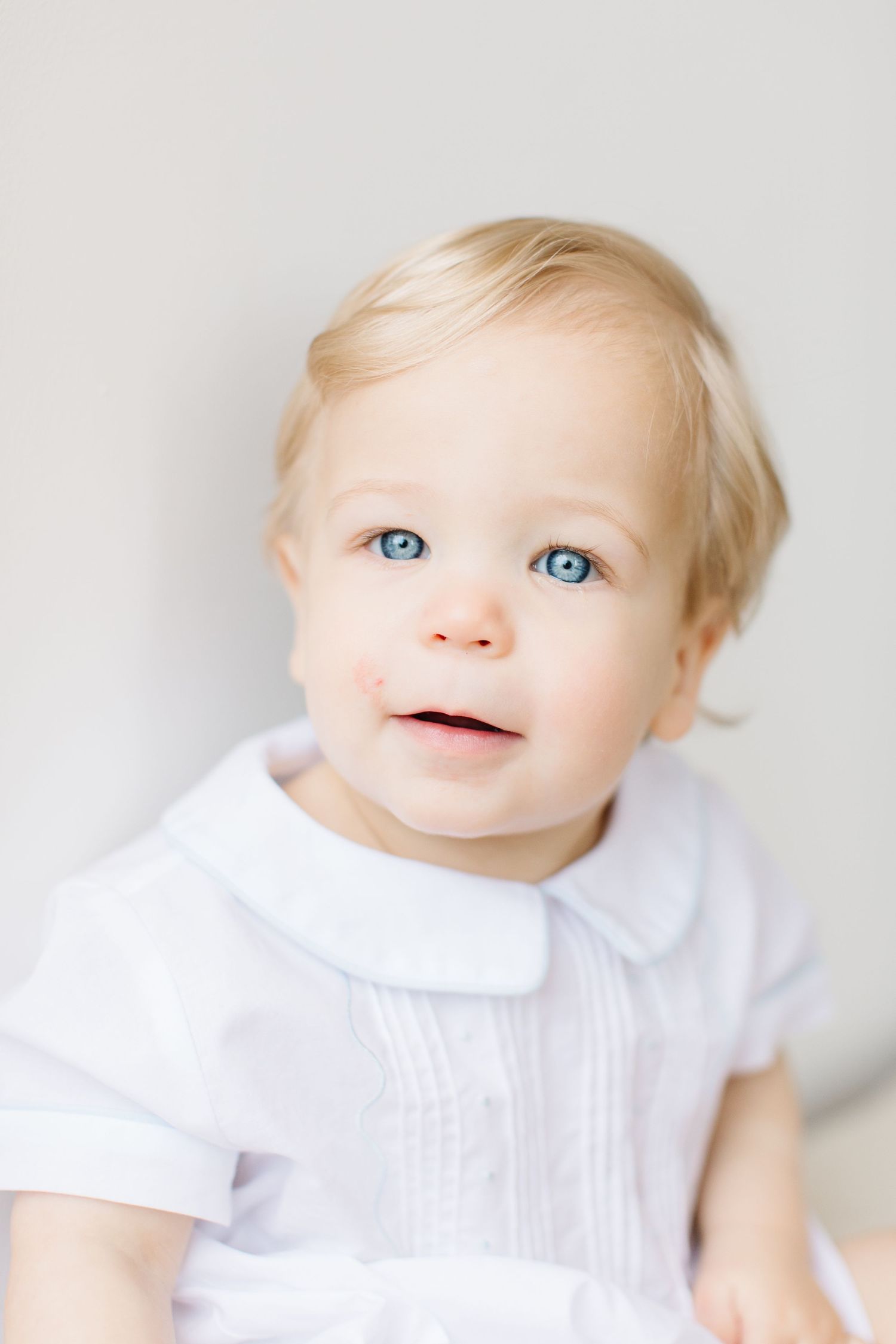 Baby wearing white collared outfit smiling and posing against light background.