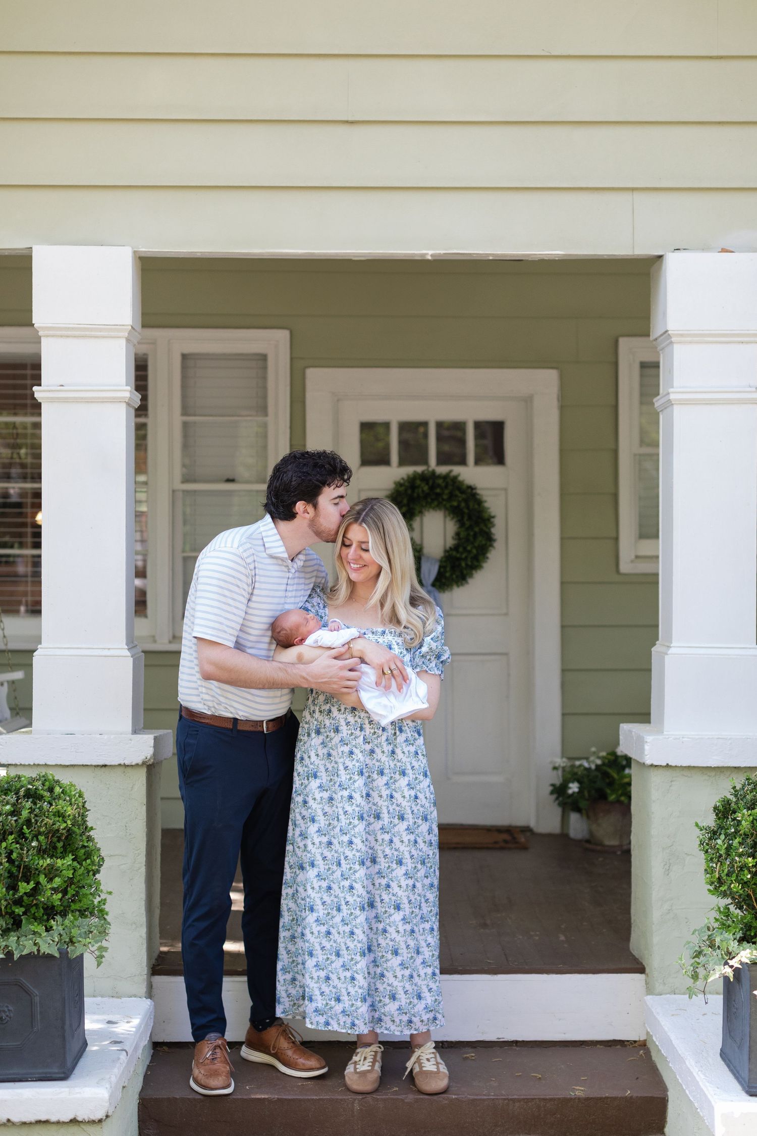 A couple stands on a porch holding their newborn baby, the woman in a floral dress and man in a striped polo.