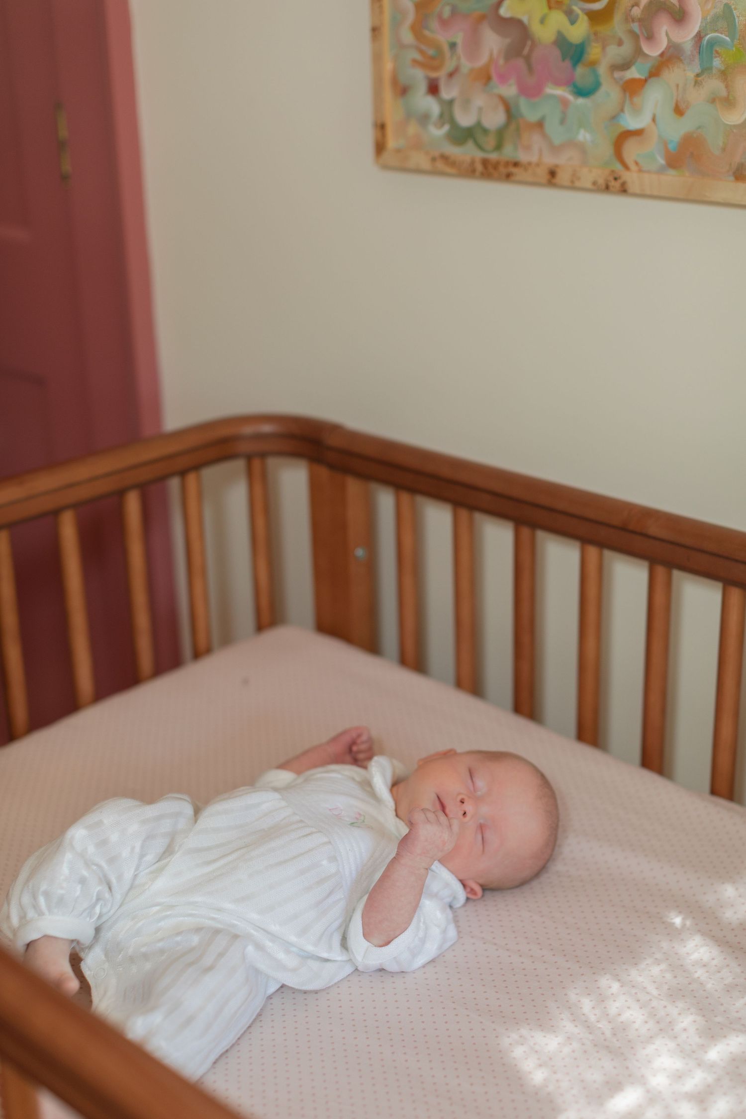 Peaceful newborn baby sleeping in wooden crib wearing white outfit in softly lit nursery.