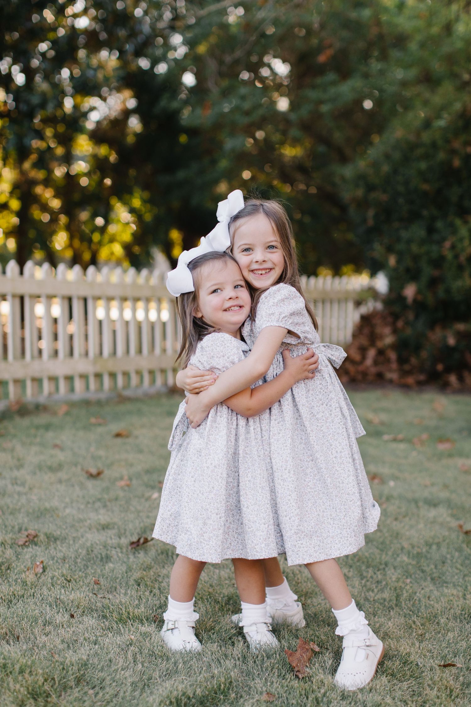 Two young children in matching white dresses and bows share sweet hugs and smiles while playing on grass near a picket fence.