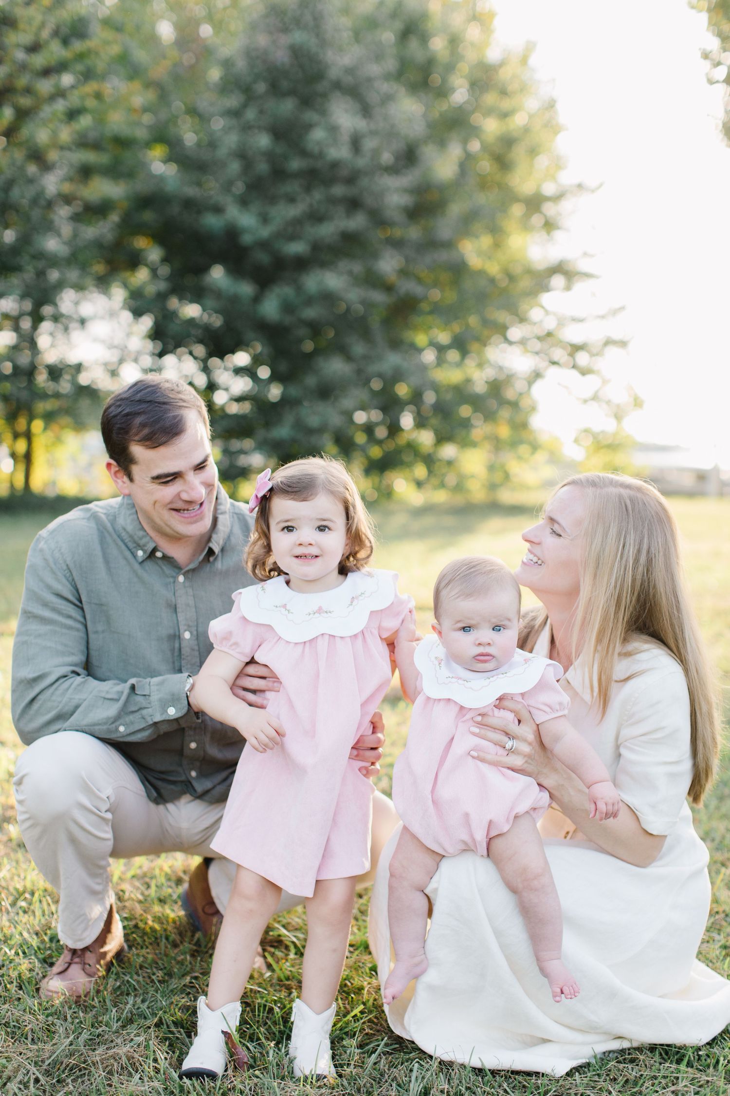 A family sits together outdoors on grass wearing coordinated pink and neutral outfits during a sunny portrait session.