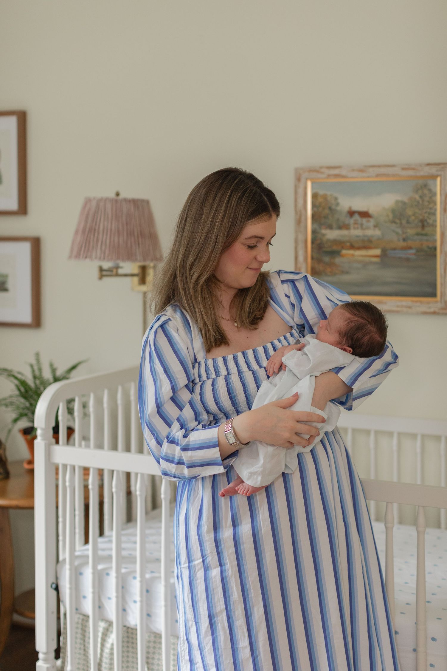 A tender moment in a nursery as a caregiver in a blue striped dress holds a sleeping newborn by a white crib.