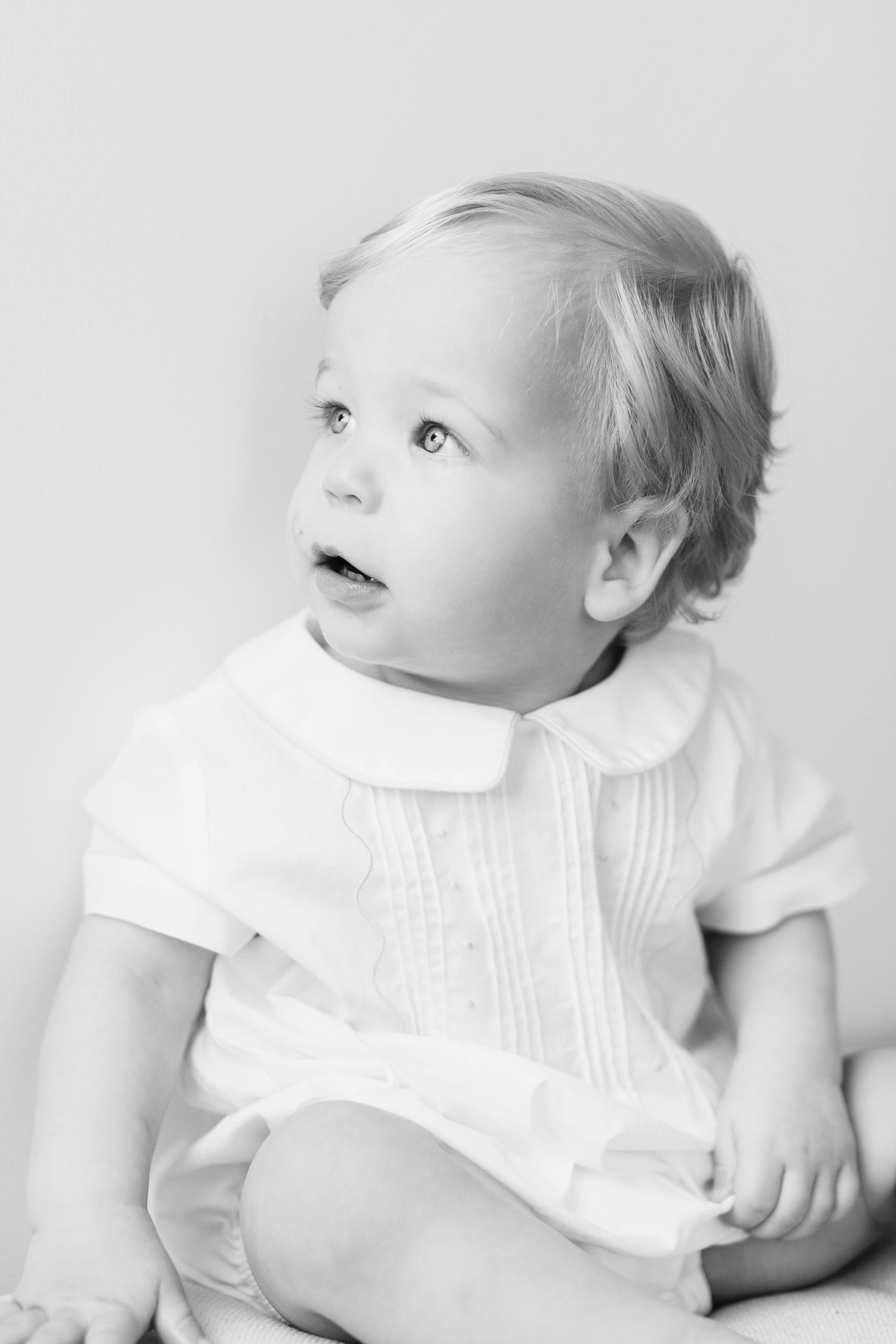 Sweet baby in white collared dress gazes upward with wonder in high-key portrait series.