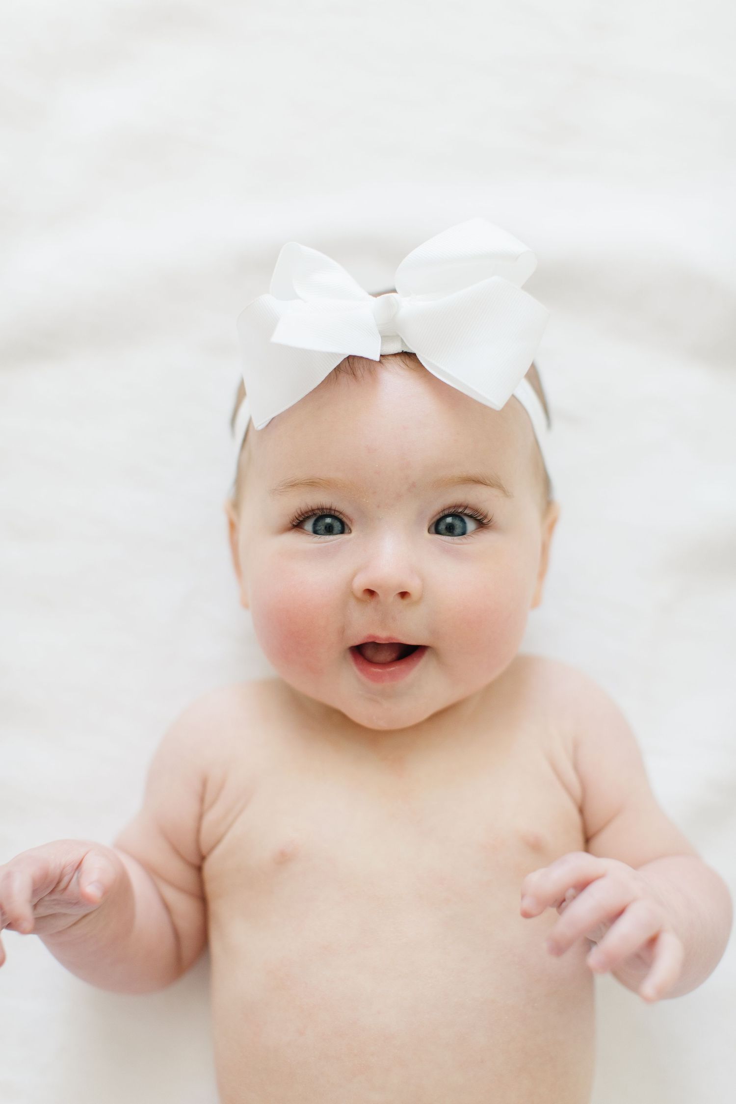 Happy baby wearing white bow headband lies on soft white bedding showing bright smile.