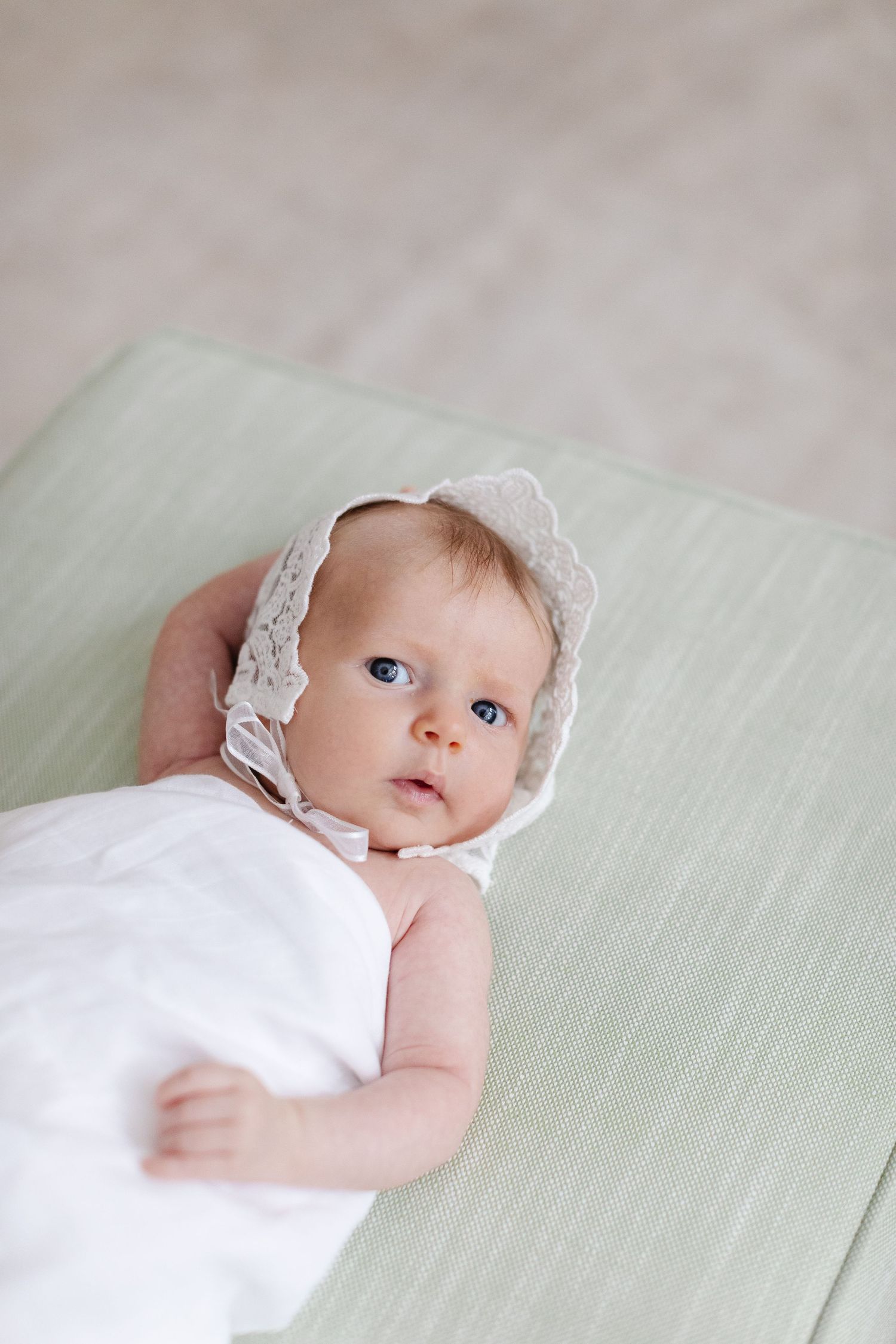 Baby wearing white lace bonnet lying on mint green surface.
