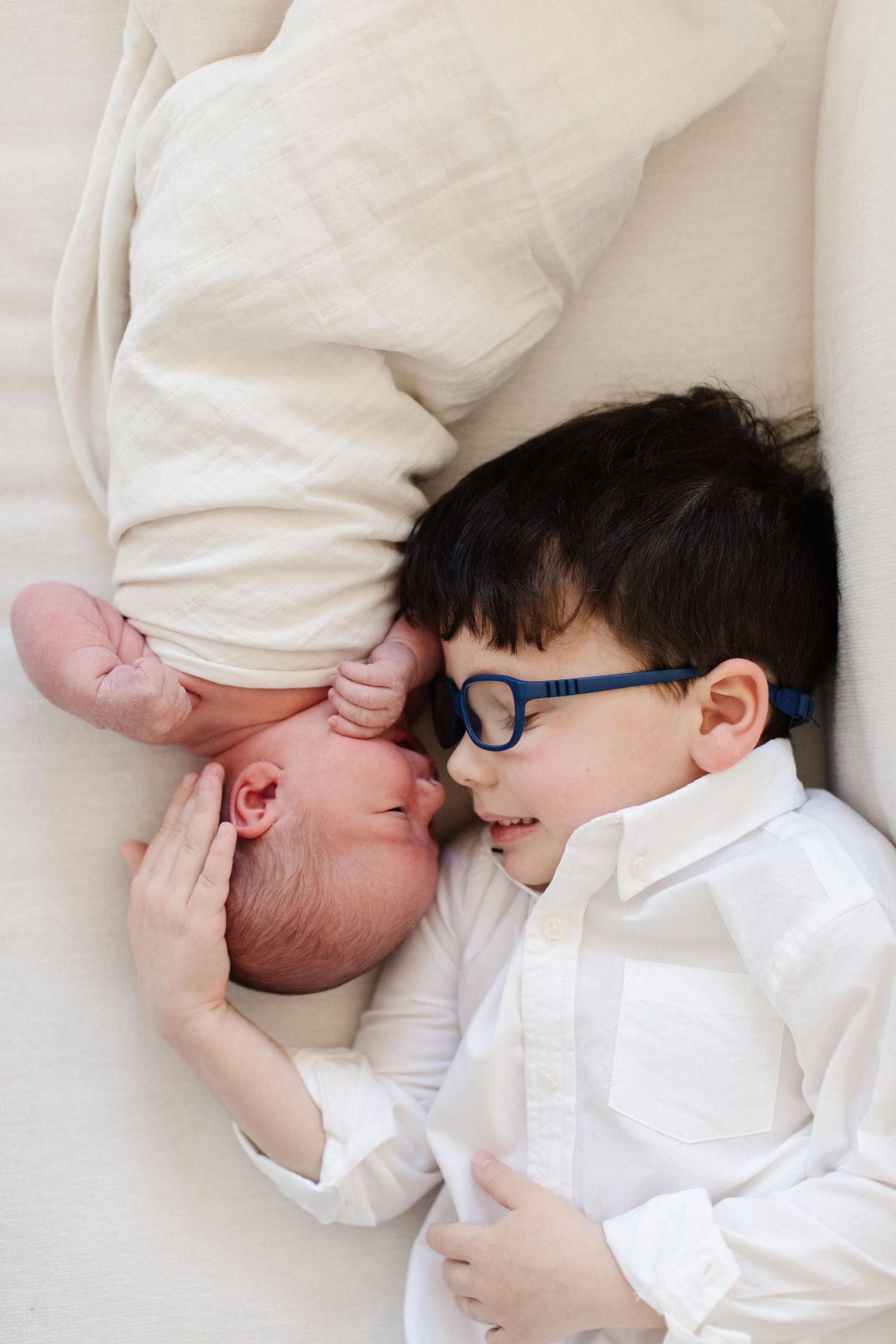 Two children in white clothing share sweet moment lying together on light bedding.