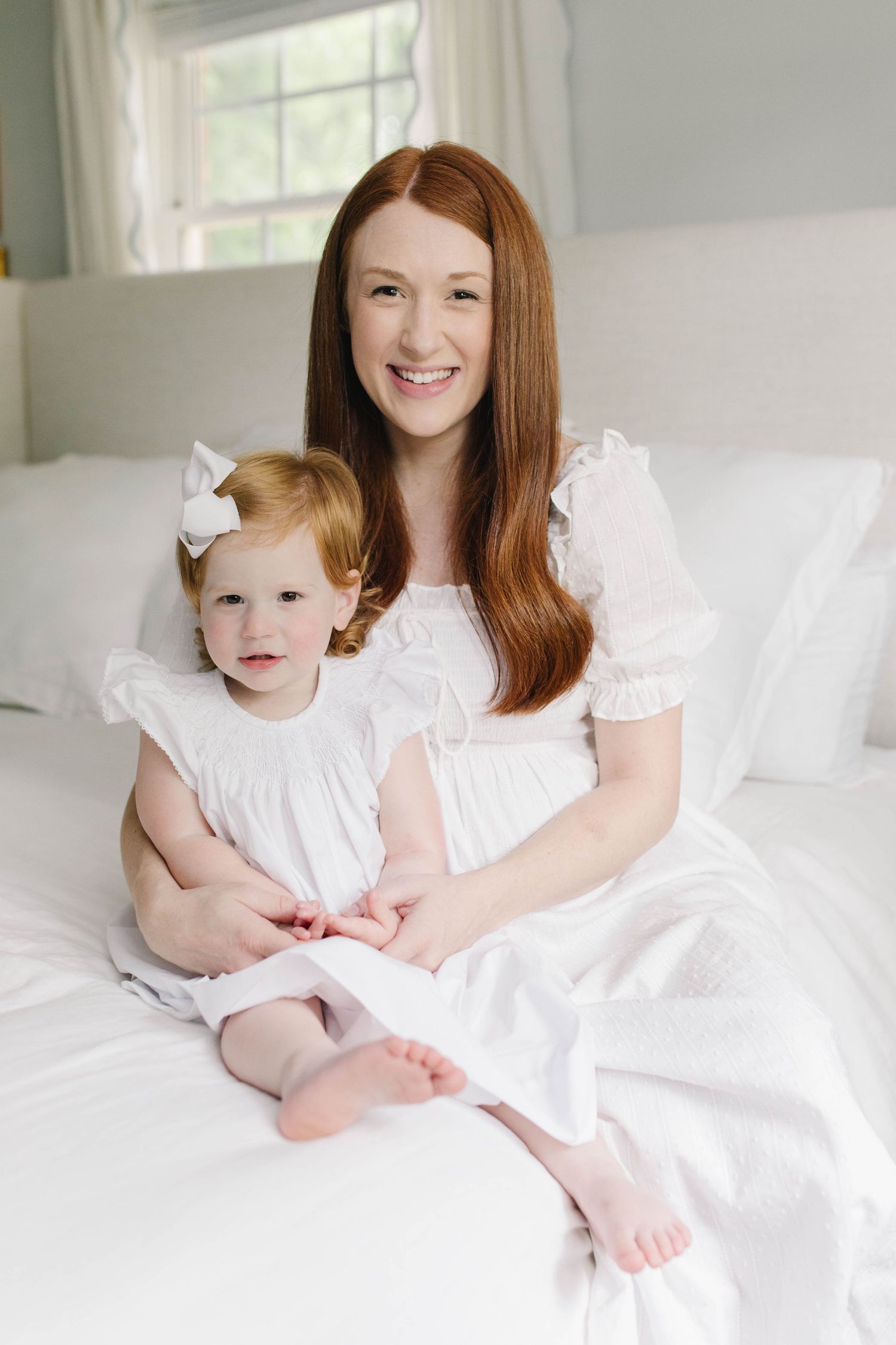 Mother and child wearing matching white dresses pose together on white bedding.