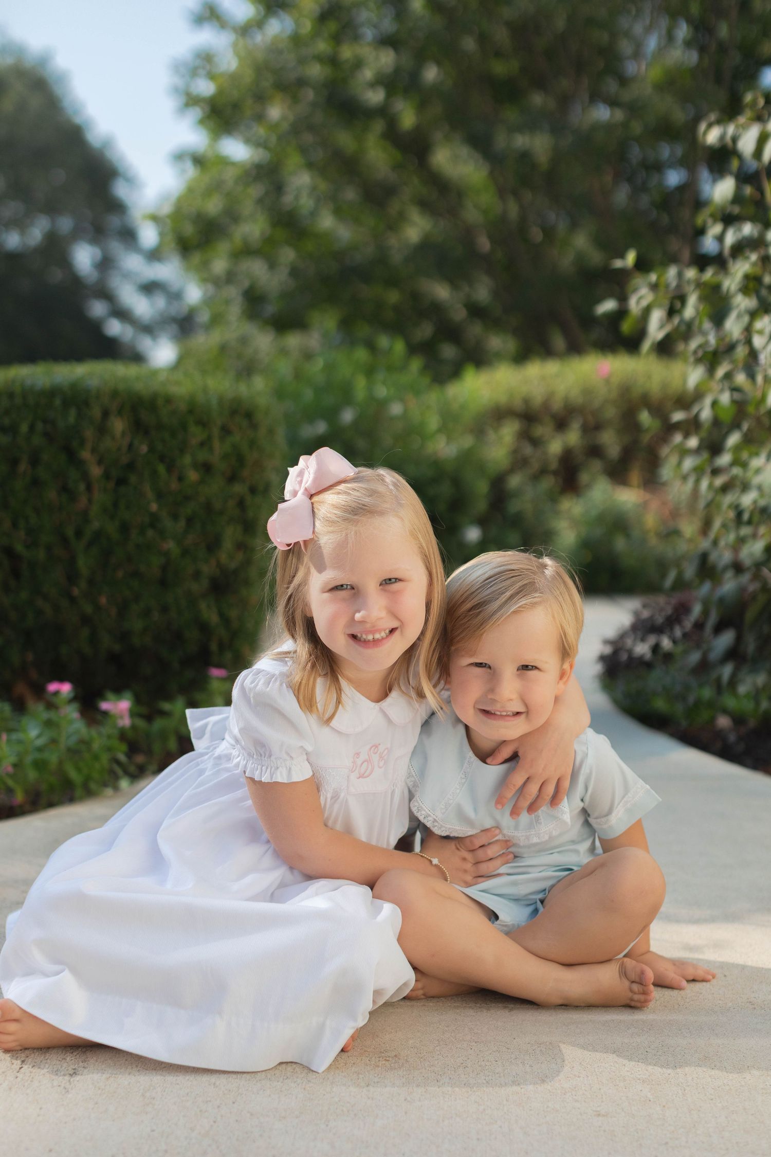 Two young children in white dresses laying together and smiling in a garden setting.