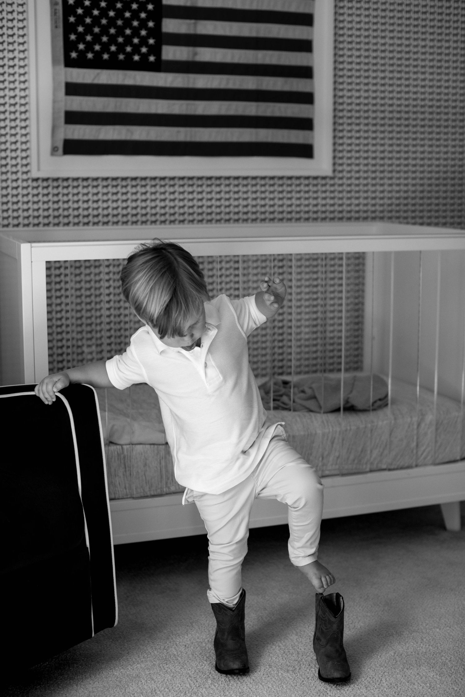 A small child stands in oversized boots beneath an American flag, wearing all white in a black and white photograph.