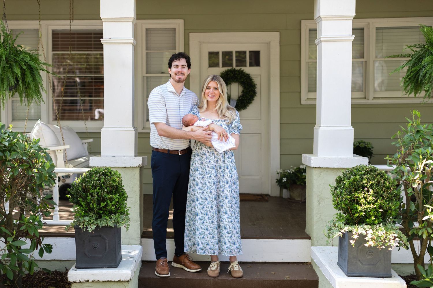 Happy couple holding newborn baby on front porch of green home with white columns and lush greenery.
