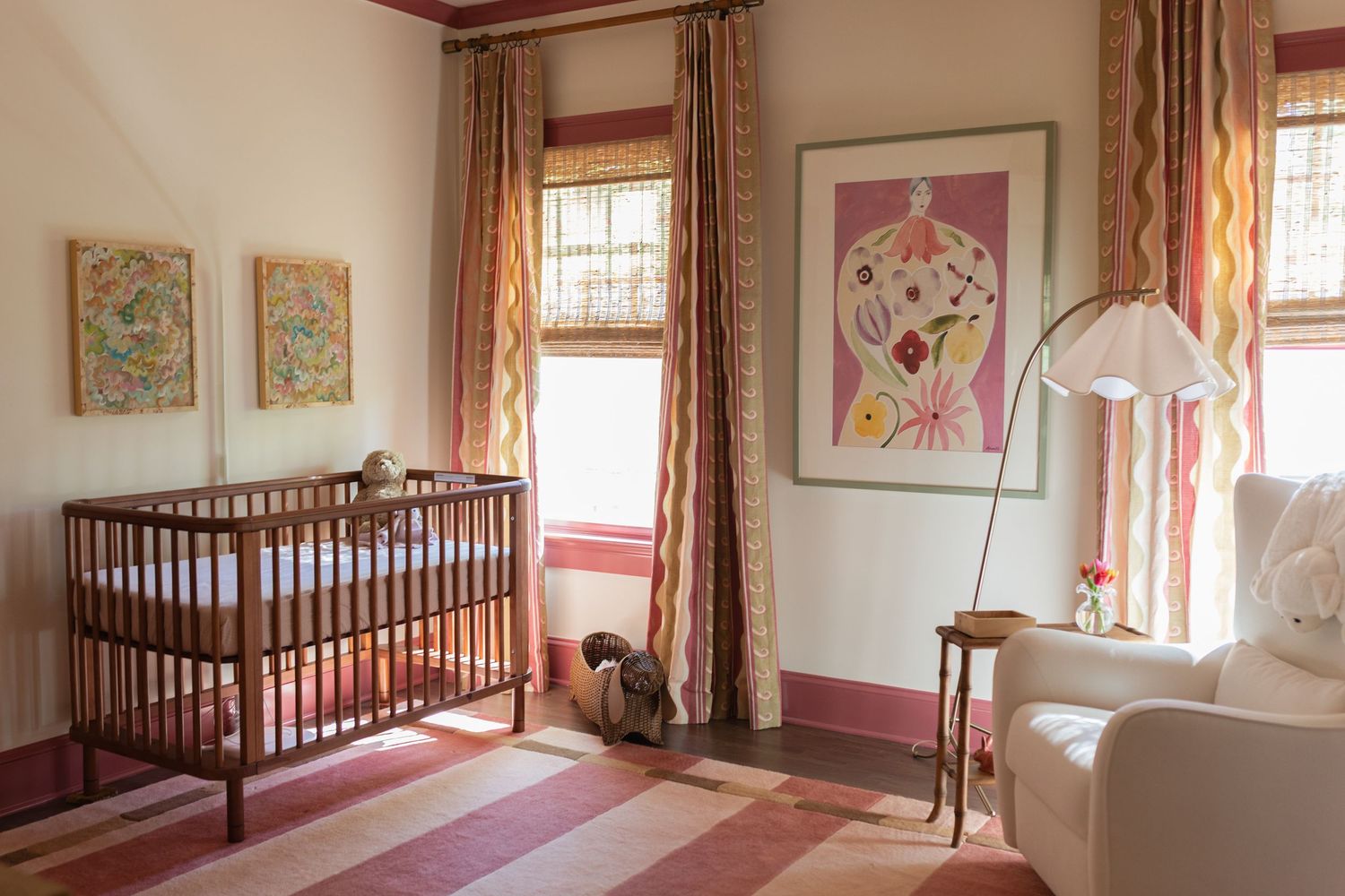 Pink and cream nursery with wooden crib, striped rug, colorful curtains, framed artwork, and a cozy armchair by the window.