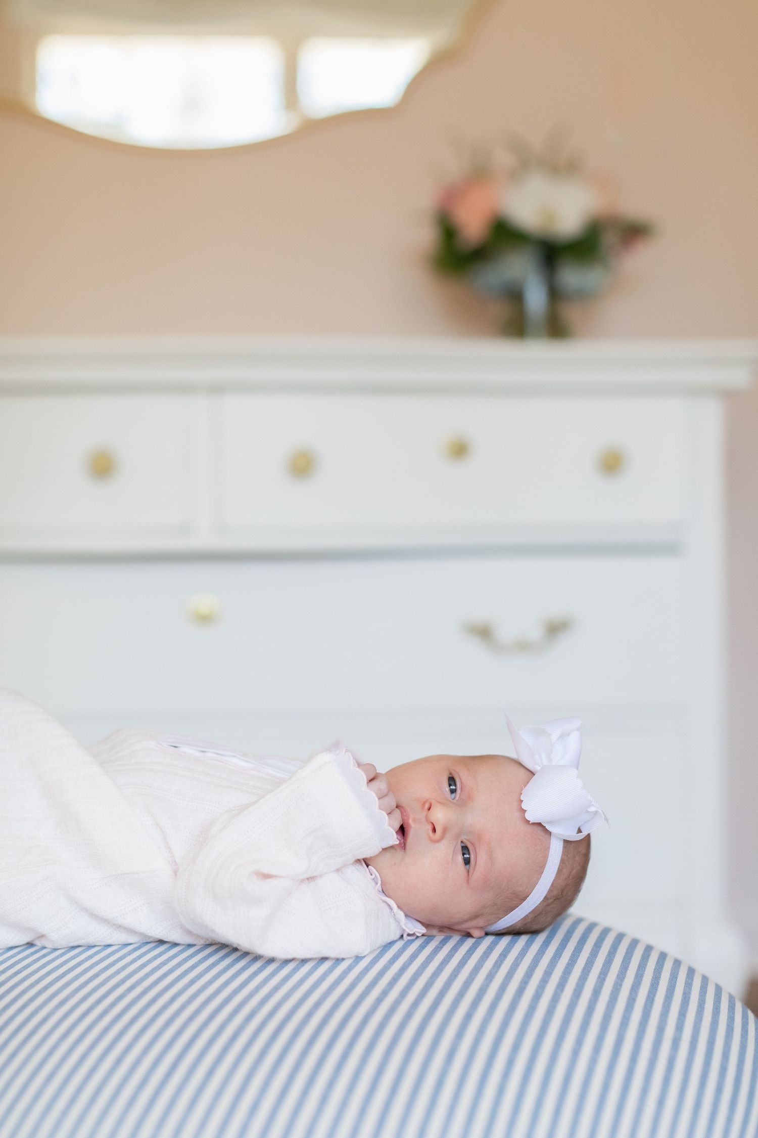 Baby wearing white bow headband and outfit rests on blue and white striped bedding.