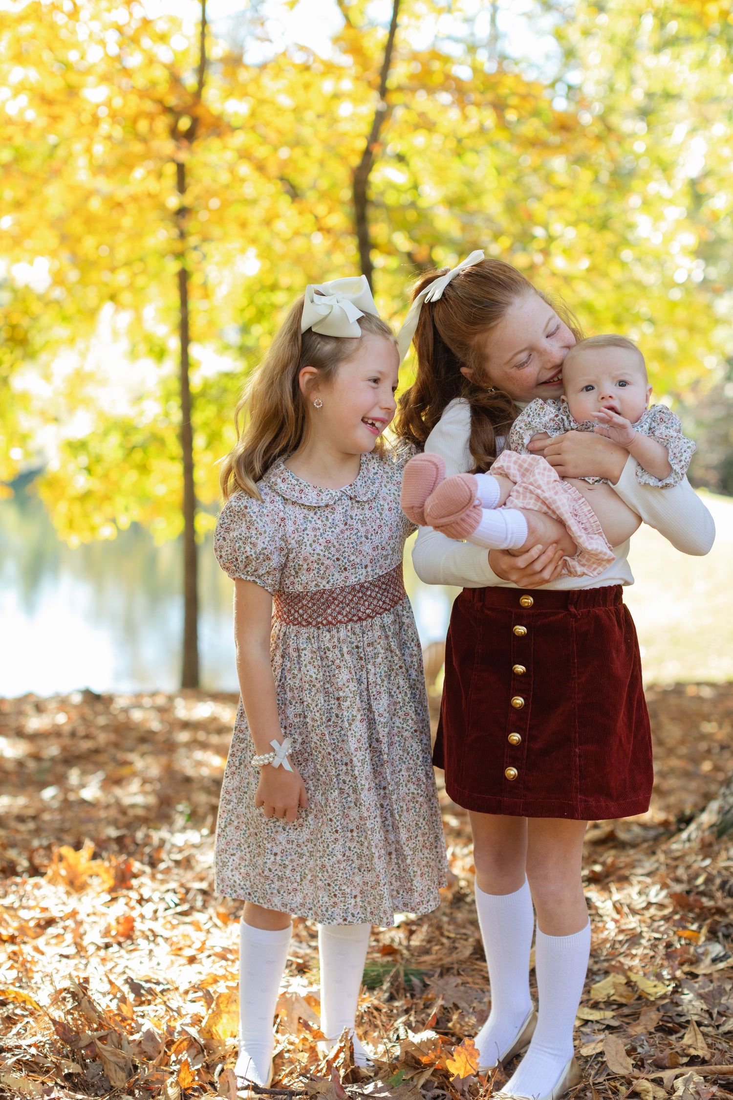 Two young girls in floral dresses and knee socks share a joyful moment with a baby among golden autumn leaves by a lake.
