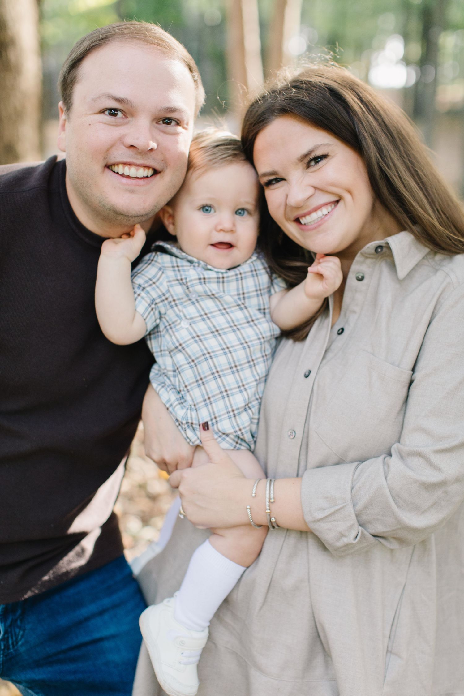 A family portrait outdoors with a young child wearing a plaid outfit and white socks between two adults.